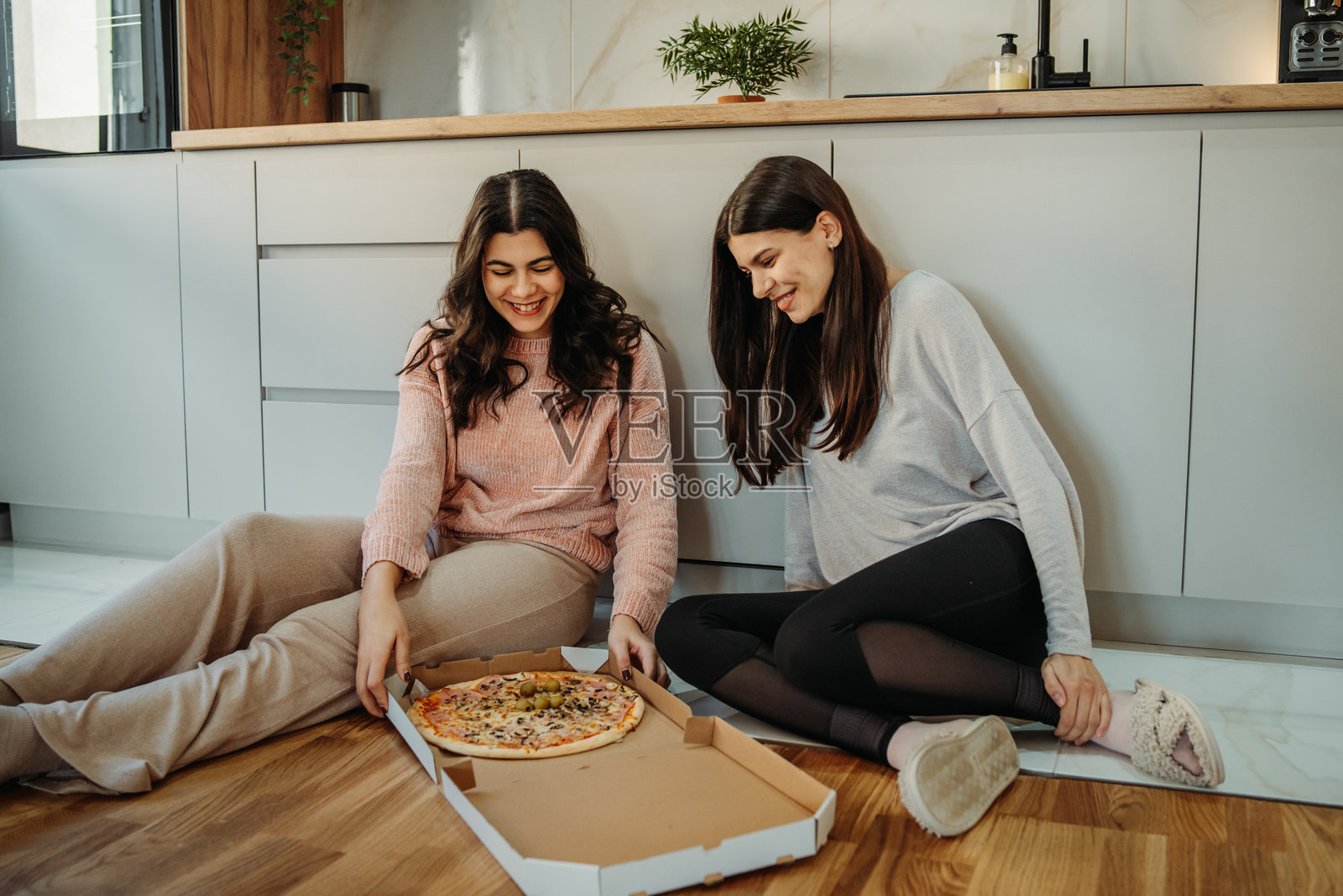 年轻的白人女性和她的姐妹坐在厨房地板上吃披萨照片摄影图片