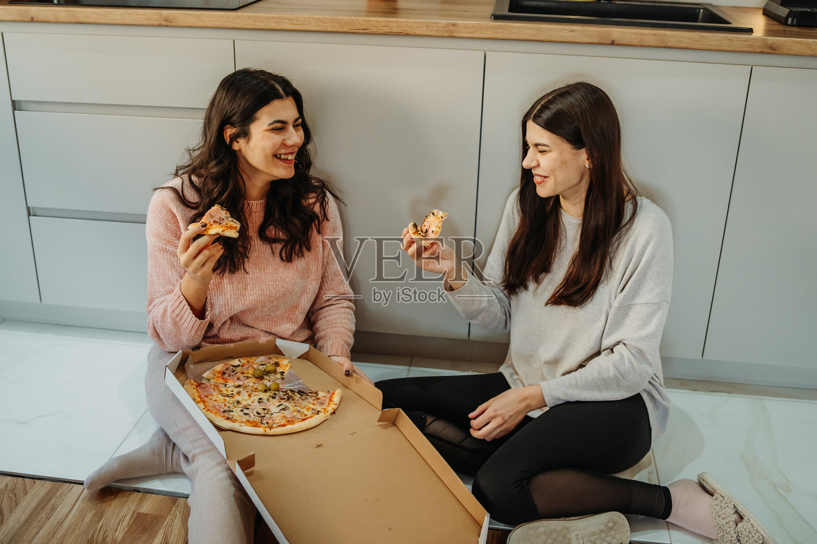 年轻的白人女性和她的姐妹坐在厨房地板上吃披萨照片摄影图片