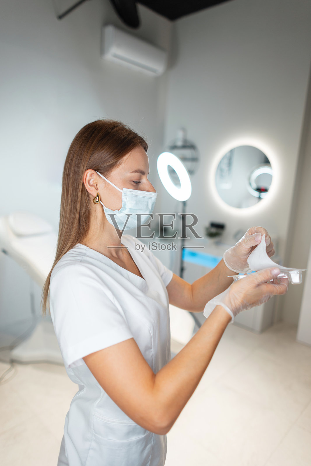 美容师女孩，穿着白大褂、戴着口罩的医生，在诊所的美容健康工作室中拆封填充剂注射器。照片摄影图片