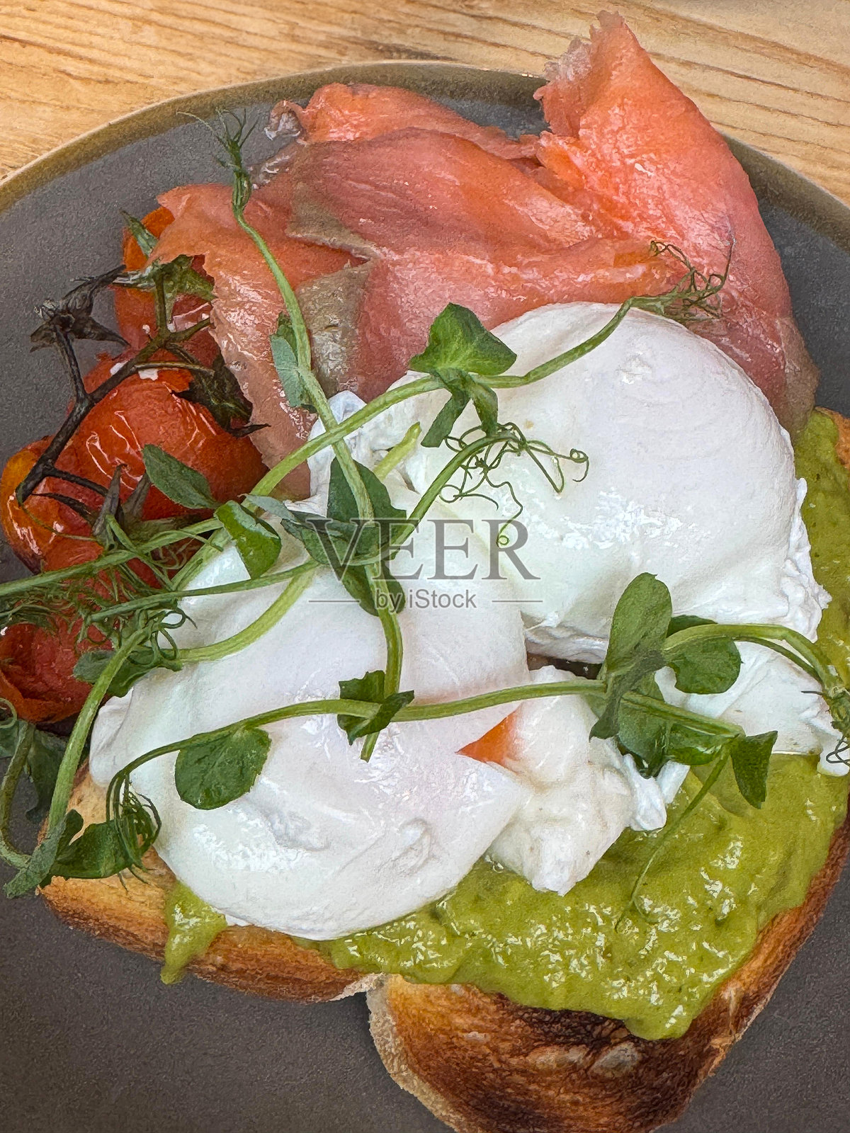 特写镜头展示了一张木桌，上面摆放着早餐：一盘白面包吐司，上面放有捣碎的鳄梨和水煮蛋，点缀着豌豆苗，旁边还有烤樱桃番茄和熏三文鱼，俯视角度。照片摄影图片