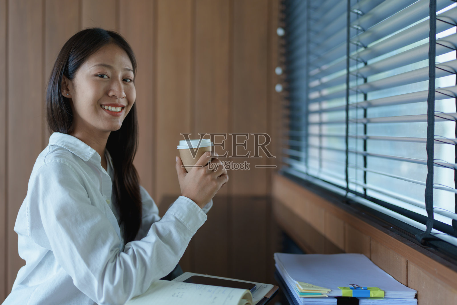 微笑的年轻女性在桌子上工作，享受咖啡，周围有笔记和透过百叶窗洒进来的自然光。照片摄影图片