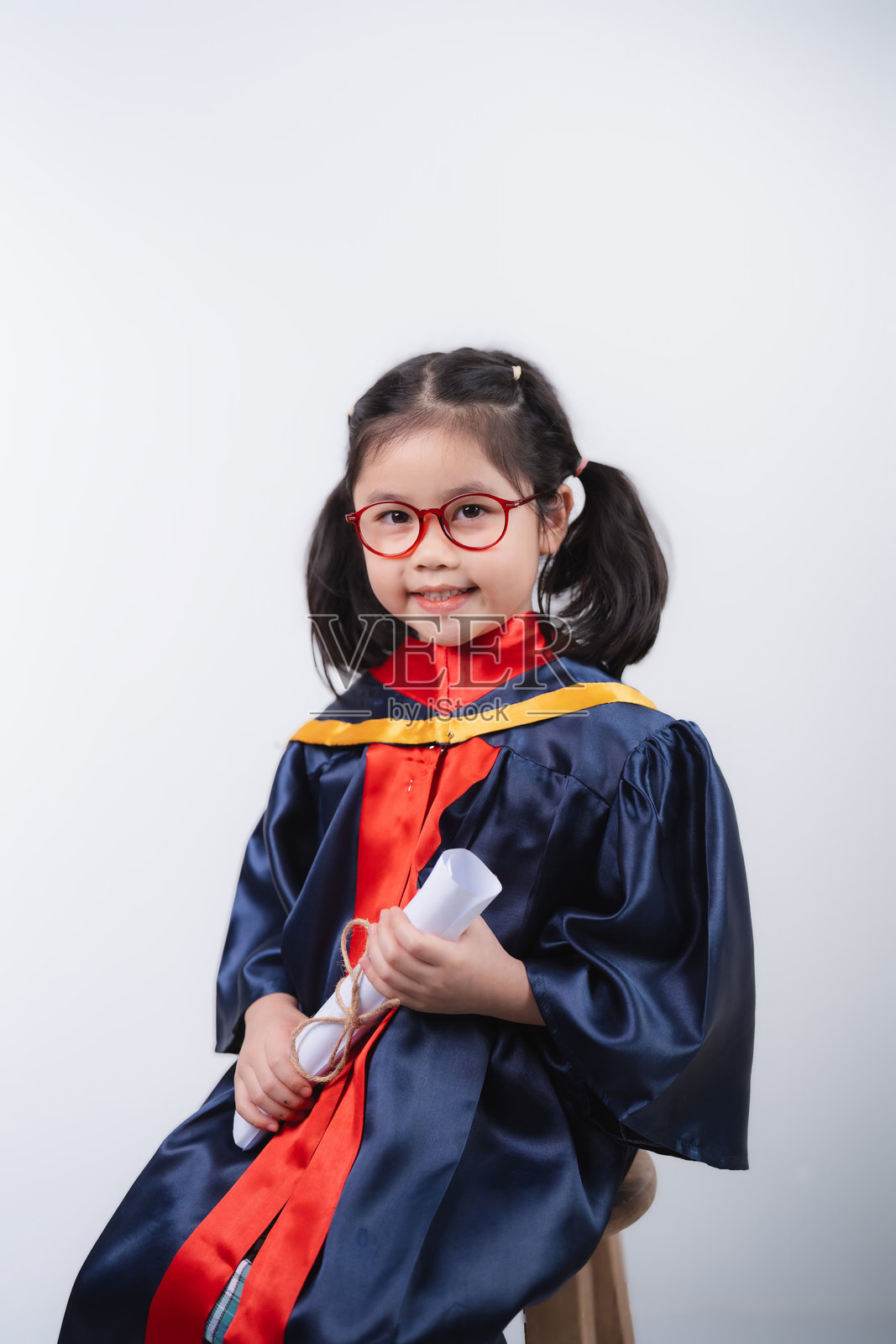 快乐的亚洲年轻女孩穿着毕业袍，手持文凭，面向镜头微笑，背景简单。照片摄影图片