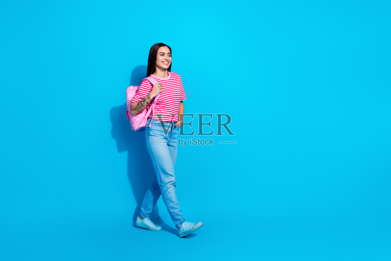 一位微笑的年轻女性，穿着粉色条纹T恤和牛仔裤，背着粉色背包，站在蓝色背景前。照片摄影图片