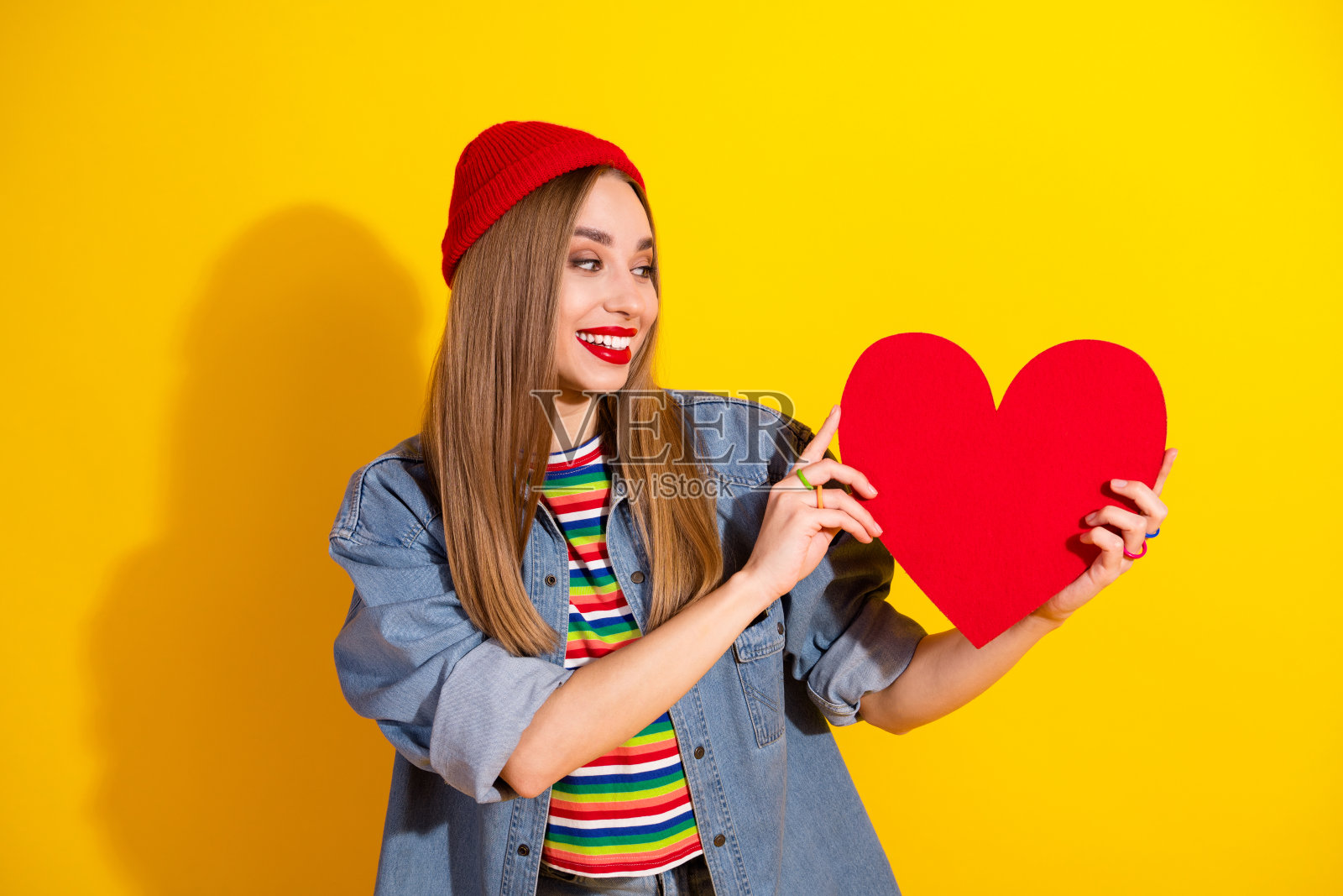 年轻女性穿着休闲服装，手持红色心形符号，背景为黄色，适合情人节和广告使用照片摄影图片