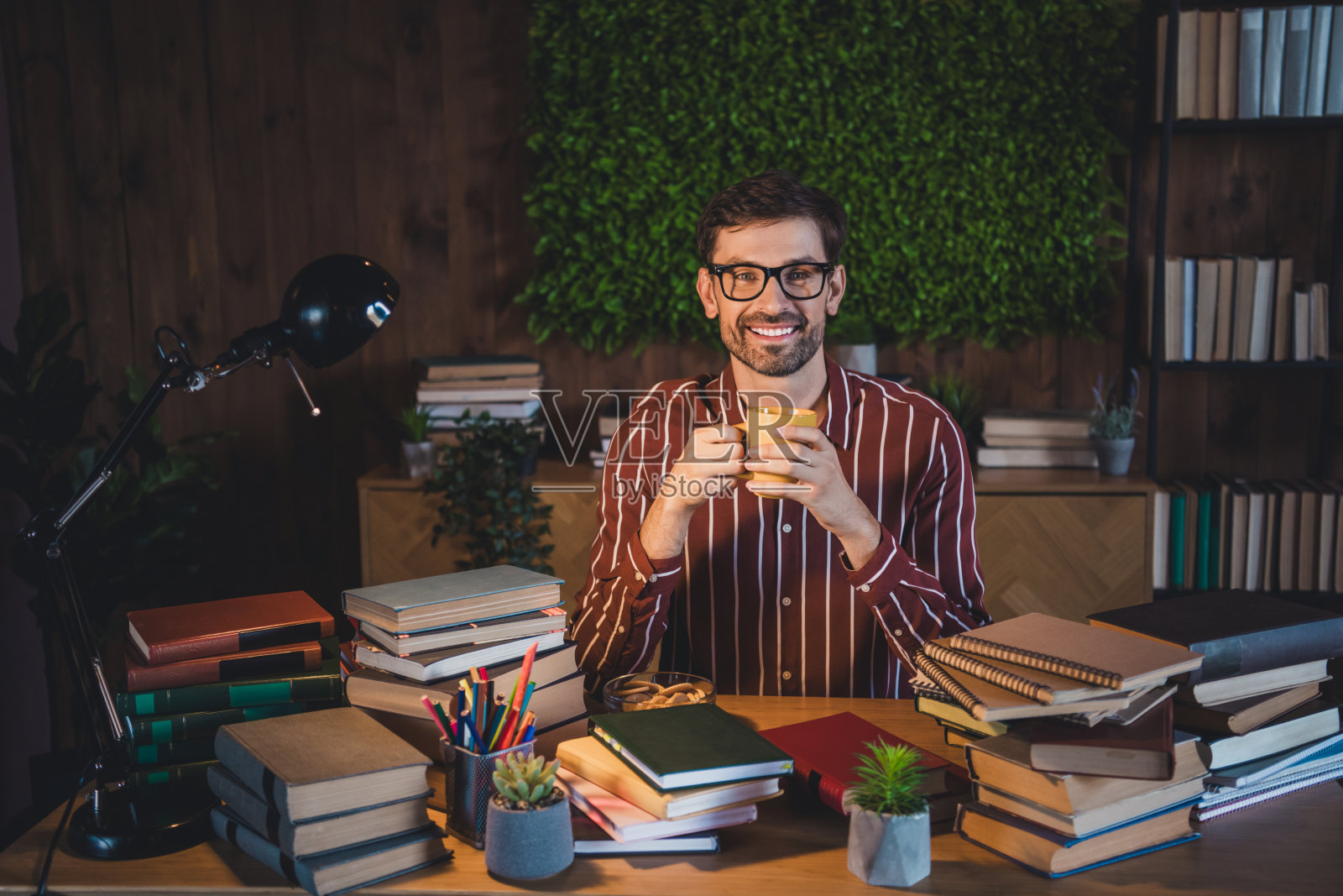 年轻男子坐在书桌前，周围堆满书籍，手里拿着一杯咖啡，微笑着享受温馨的室内环境照片摄影图片