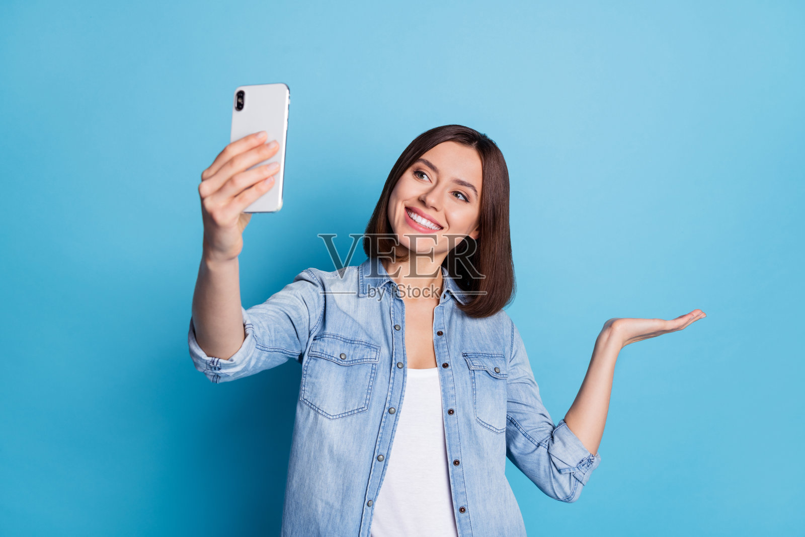 可爱的女士穿着牛仔衬衫，手持自拍设备，背景为空旷的蓝色。照片摄影图片