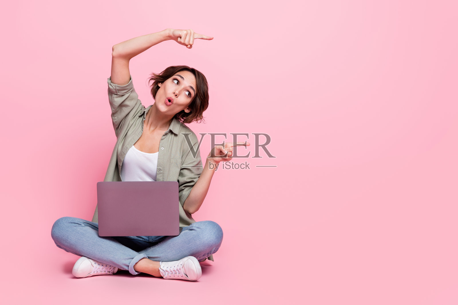 惊讶的女士全身照，撅嘴直视空无一物的手指，背景为粉色。照片摄影图片