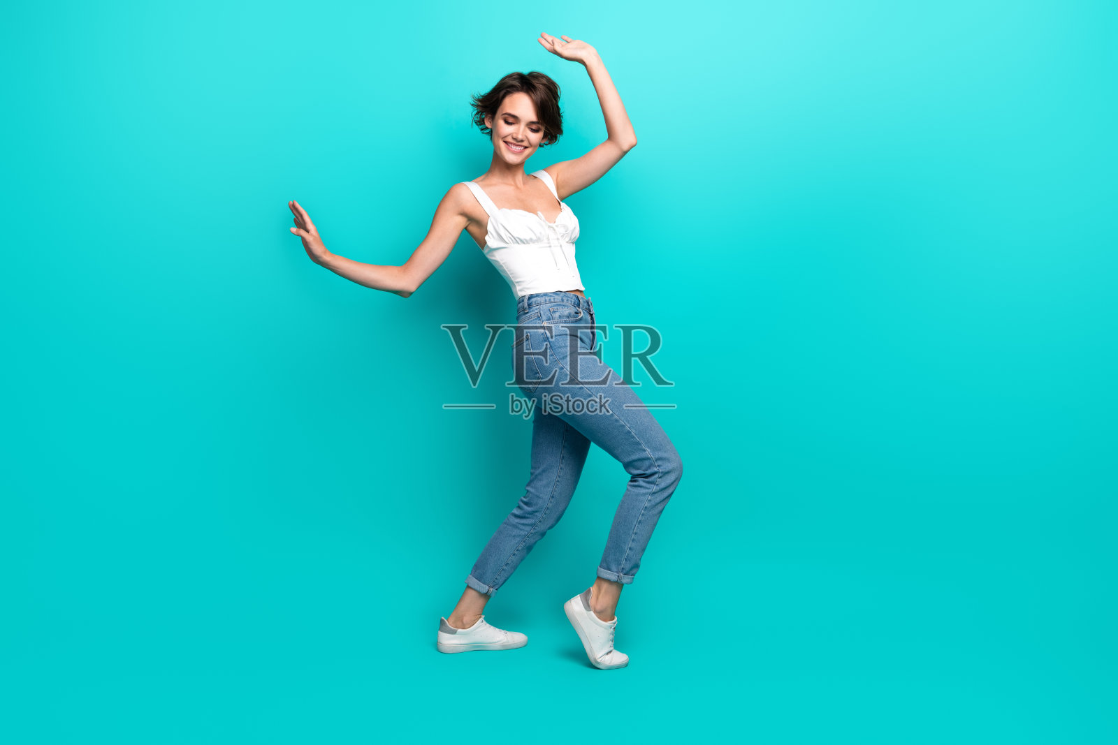 微笑的年轻女性穿着时尚休闲的牛仔裤和白色上衣，在充满活力的青绿色背景下快乐地跳舞，展现出魅力和积极的态度。照片摄影图片