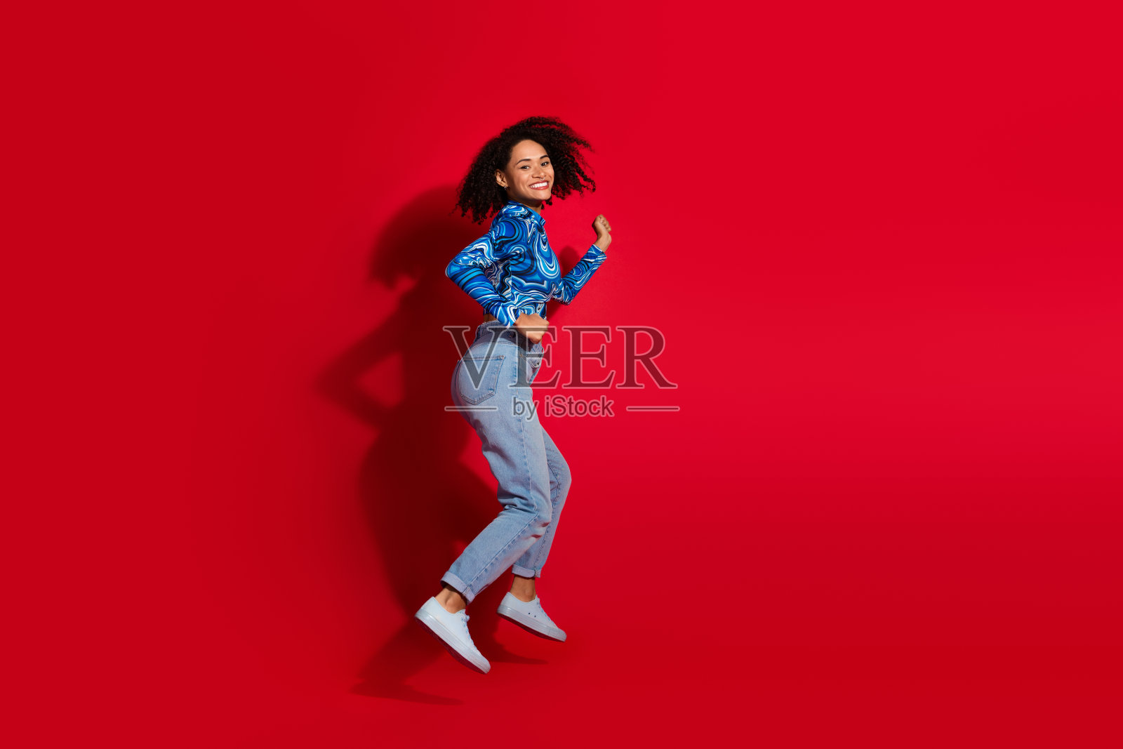 充满活力的女性在鲜艳的红色背景下愉快地跳舞，穿着时尚的蓝色印花上衣和休闲牛仔裤，面带灿烂的笑容。照片摄影图片