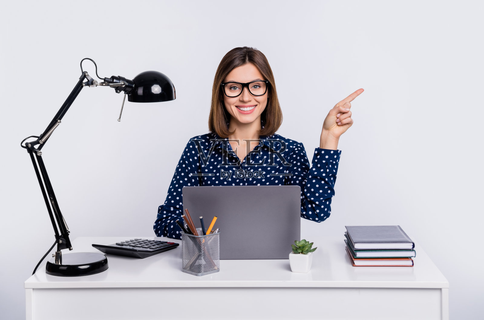 自信的女士坐在桌子旁，手指指向空白处，穿着点点衬衫，背景为灰色。照片摄影图片