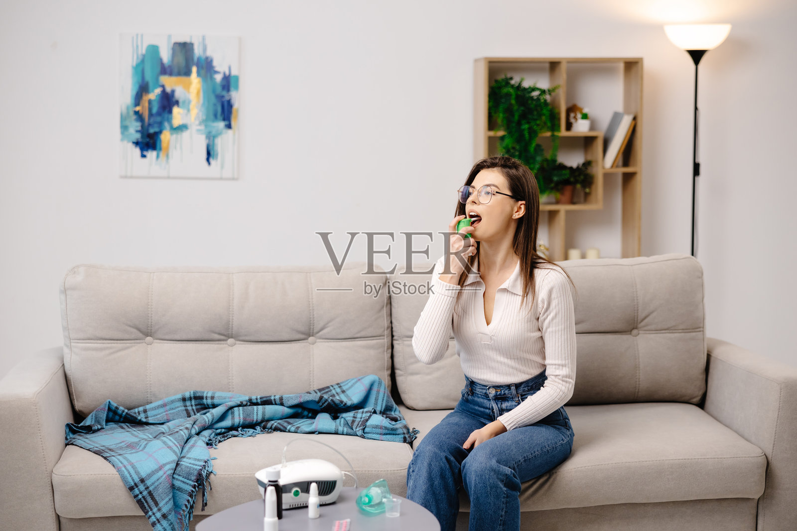 年轻女性穿着休闲服，使用止痛喷雾缓解喉咙不适，坐在家居室内的沙发上照片摄影图片