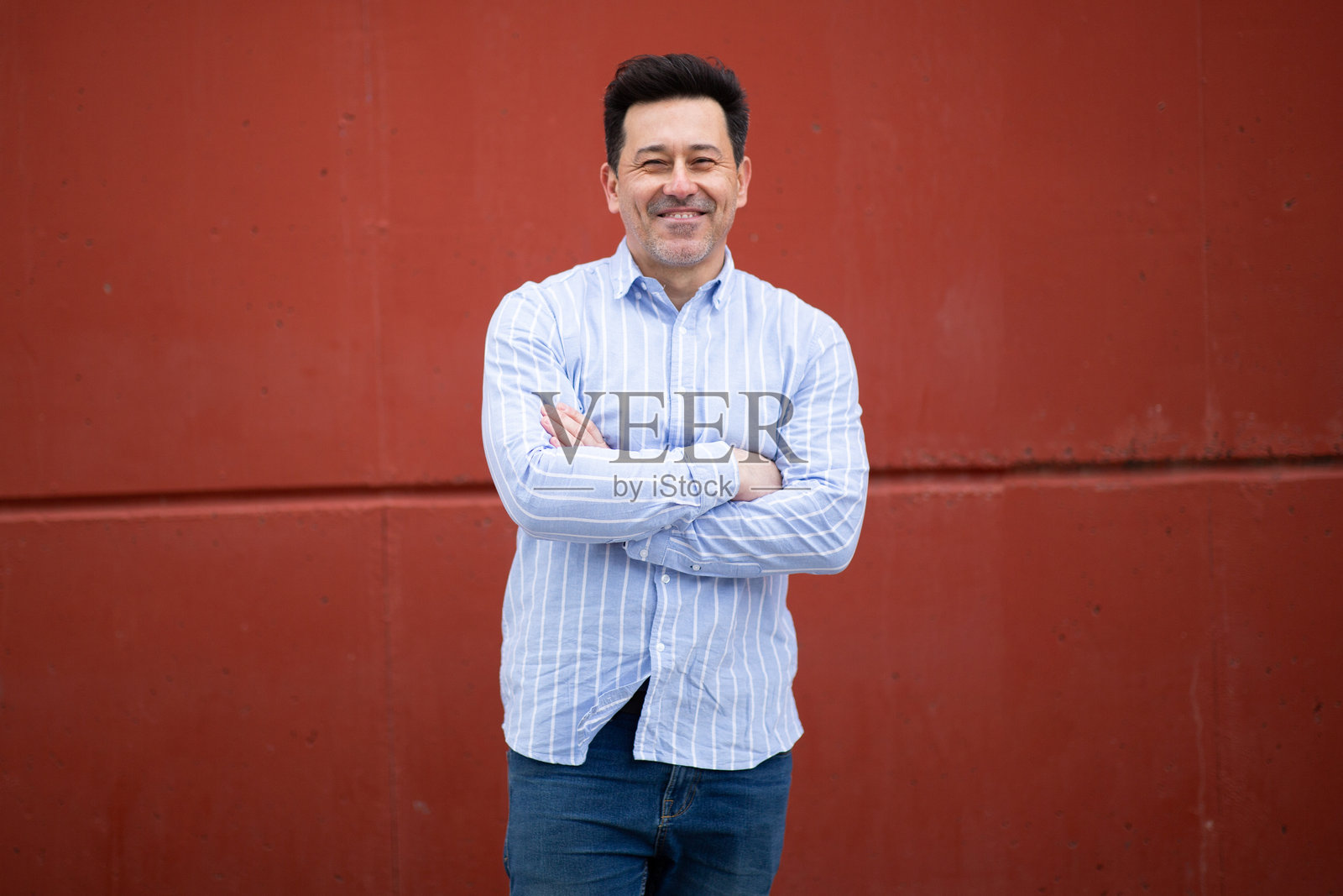 自信的成熟拉丁男性双臂交叉，站在红色墙壁前照片摄影图片