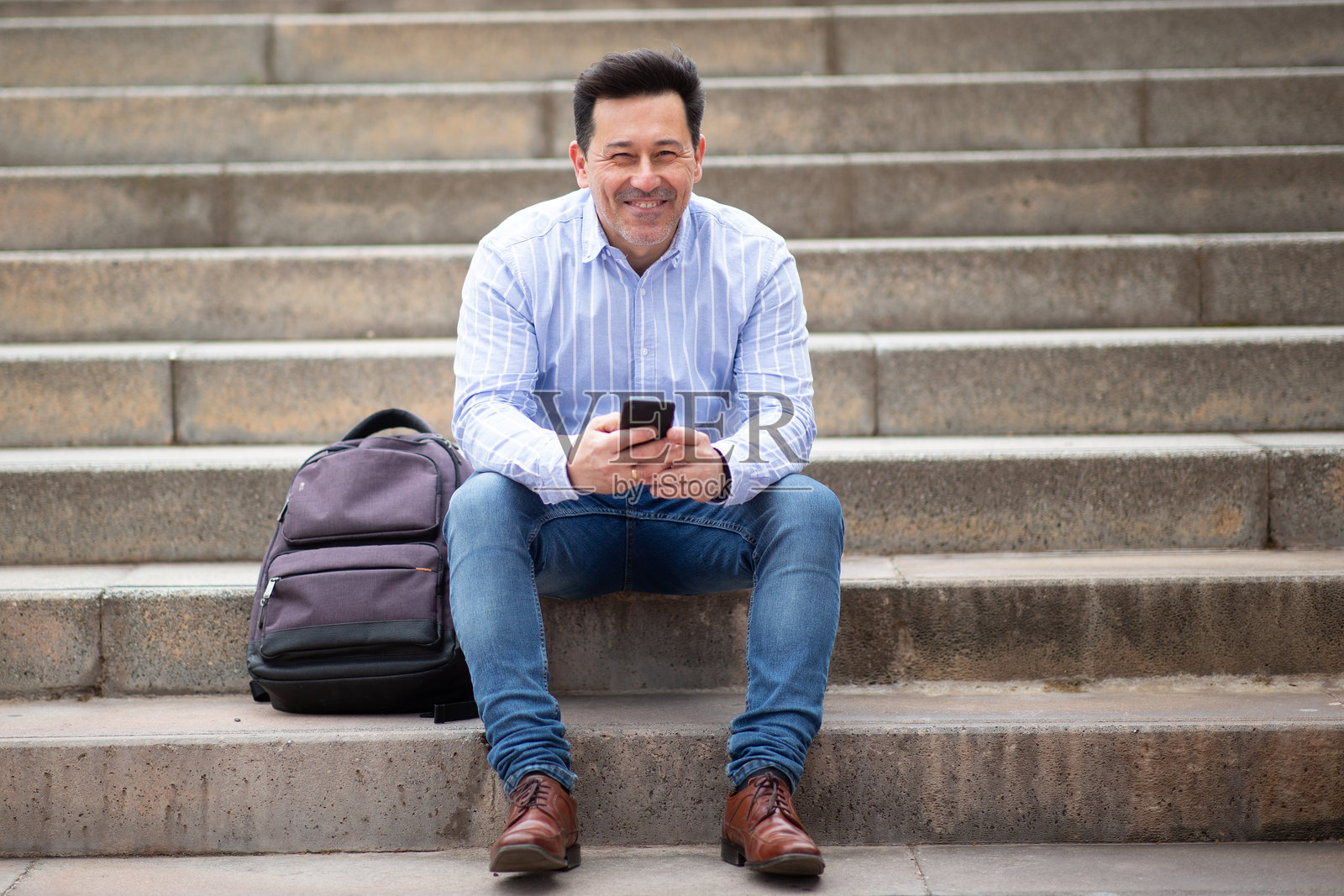 快乐的成熟拉丁男性坐在户外的台阶上，手里拿着包和手机照片摄影图片