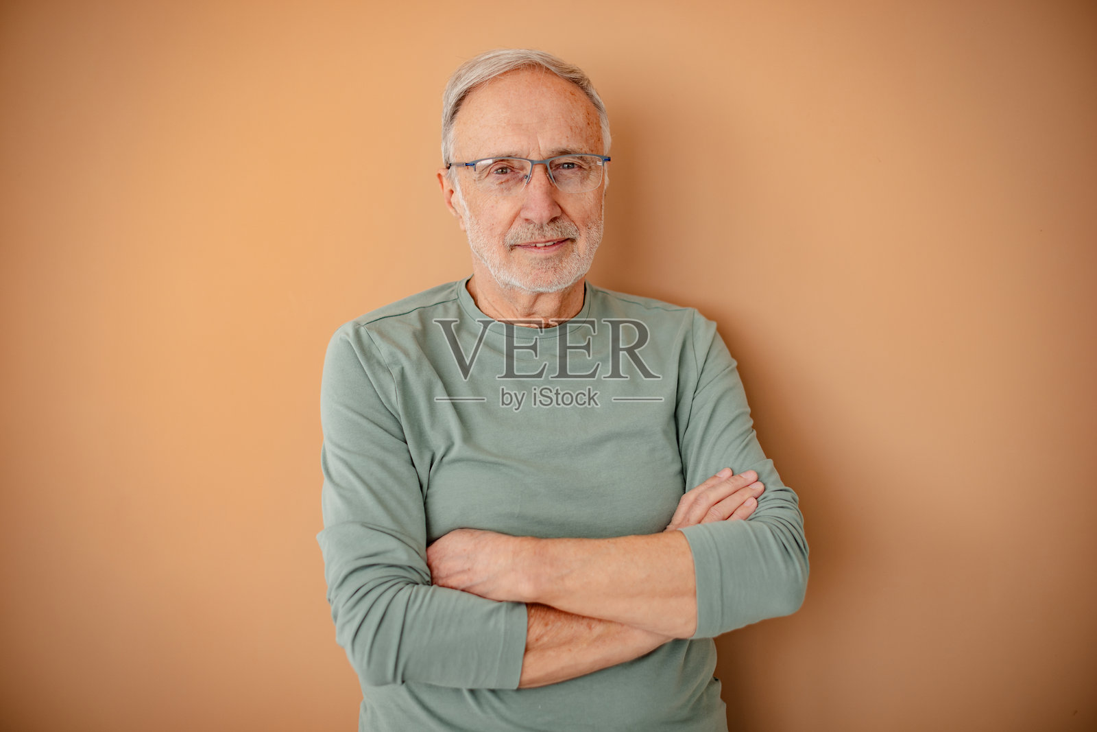 一位佩戴现代眼镜的老年人微笑着，站在温暖色调的墙壁前，身处一个休闲的室内环境中。照片摄影图片