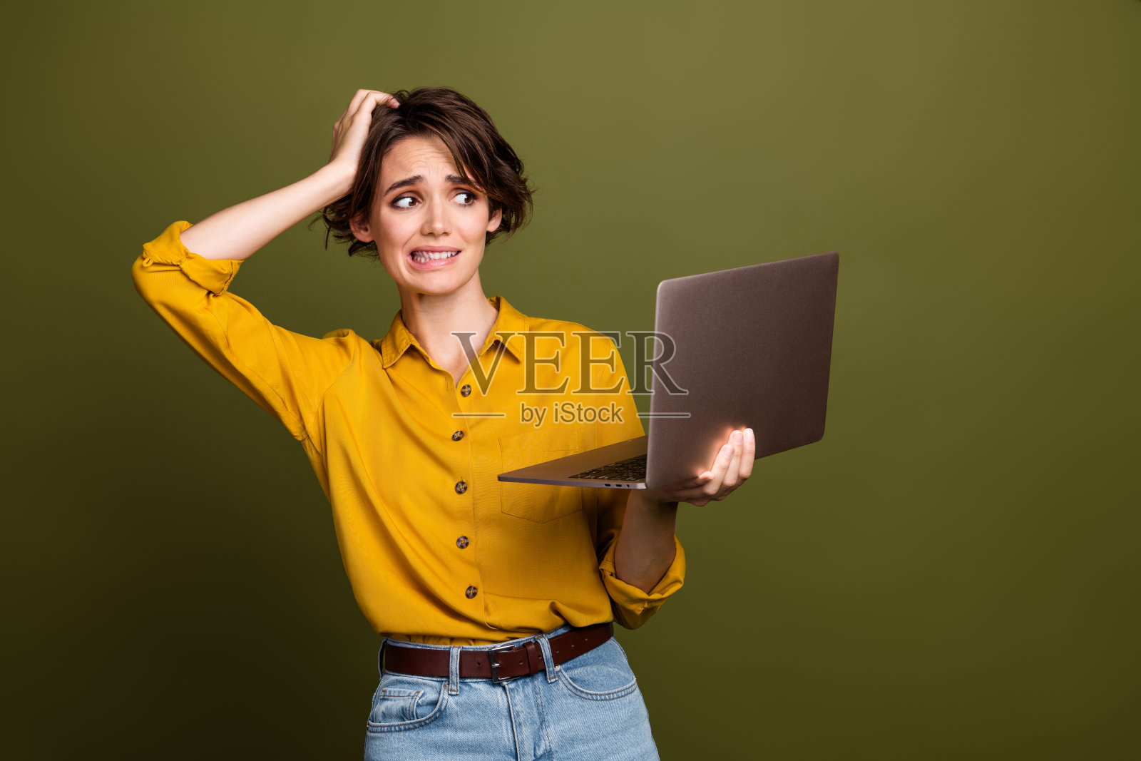 困惑的职业女性手持笔记本电脑，穿着休闲的黄色衬衫和牛仔裤，站在卡其绿色背景前。照片摄影图片