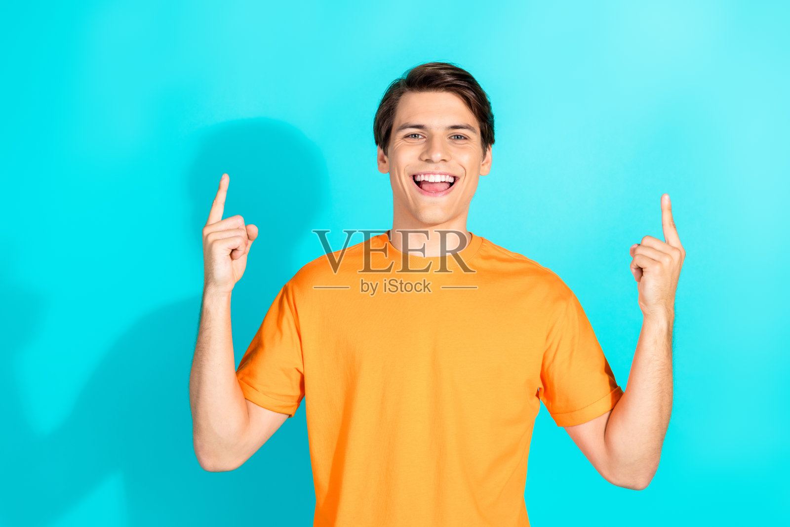 穿着橙色T恤的年轻男子在青色背景前指向上方，表达快乐和积极的情绪照片摄影图片