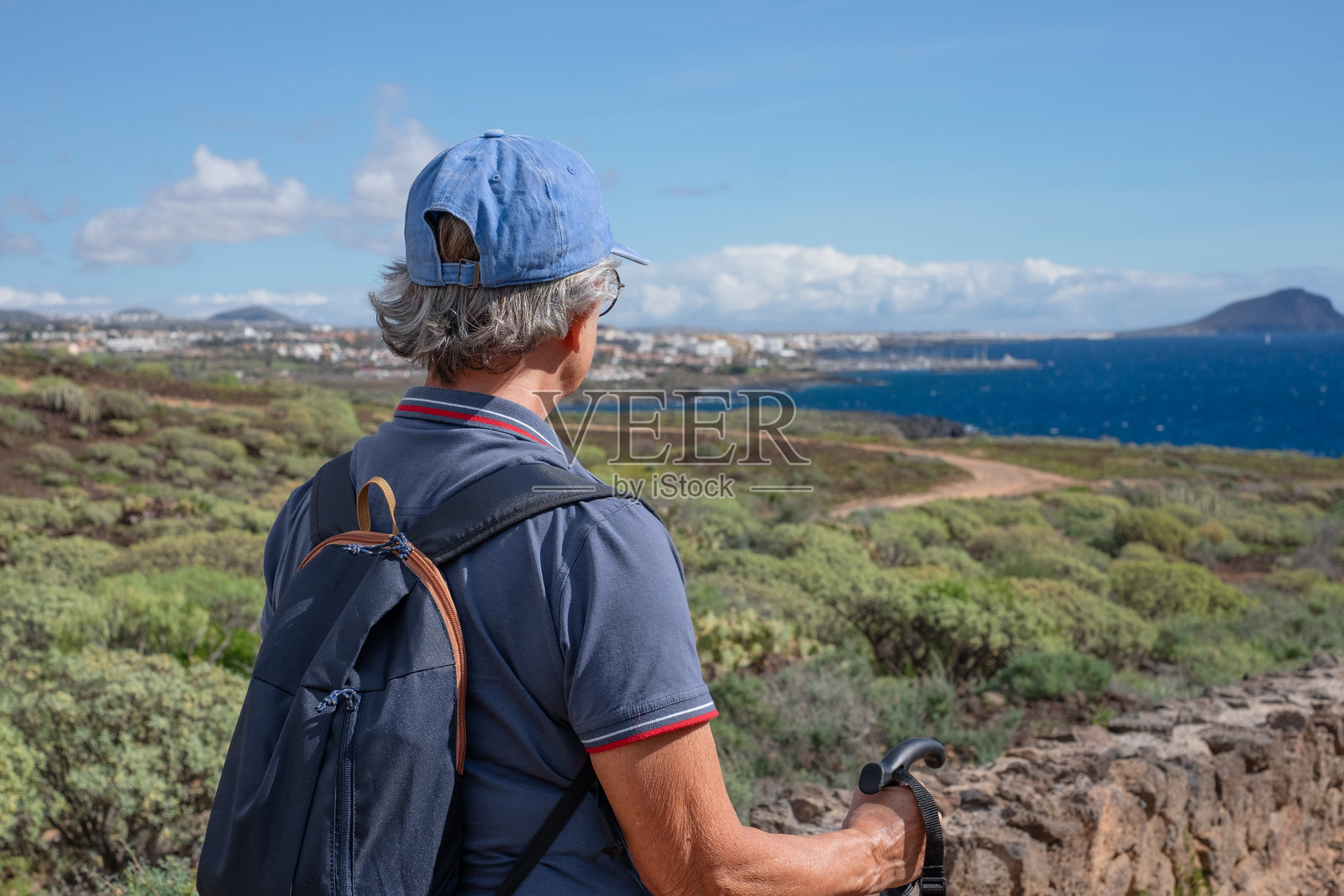 背影的老年女性背着背包在乡村户外远足，俯瞰海景。放松的白人女性享受退休生活，过着健康的生活方式，水面上的地平线。照片摄影图片