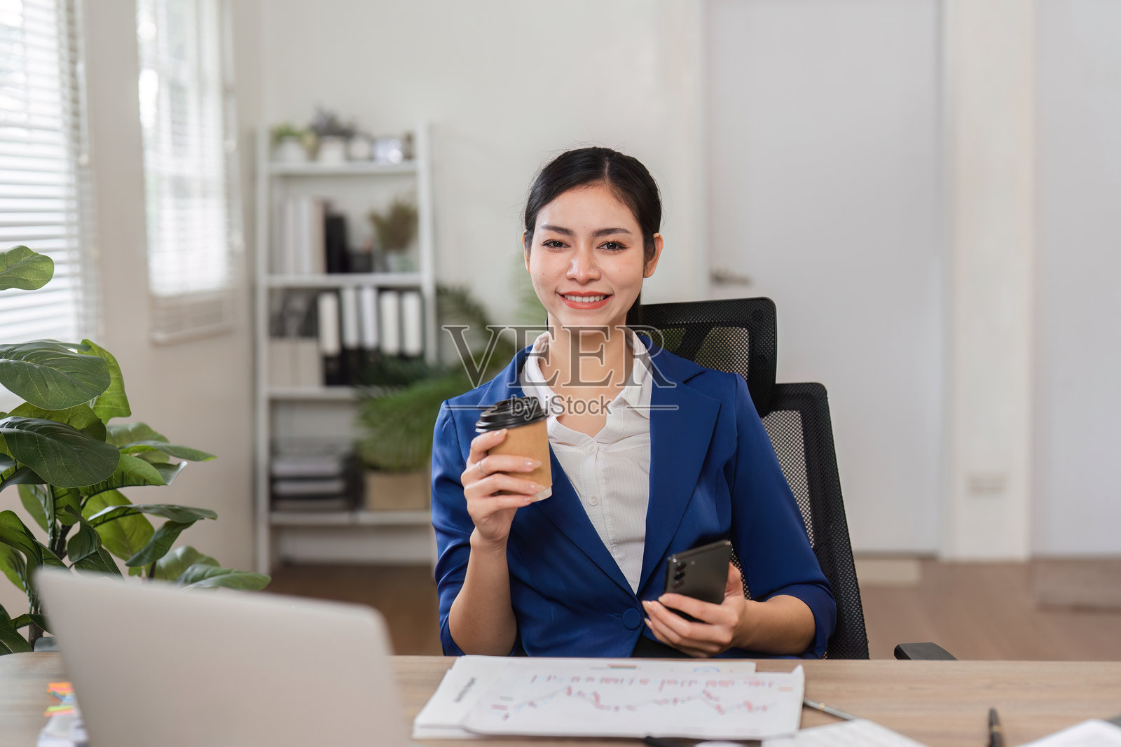 微笑的女商人在办公室里喝着咖啡，使用智能手机。照片摄影图片