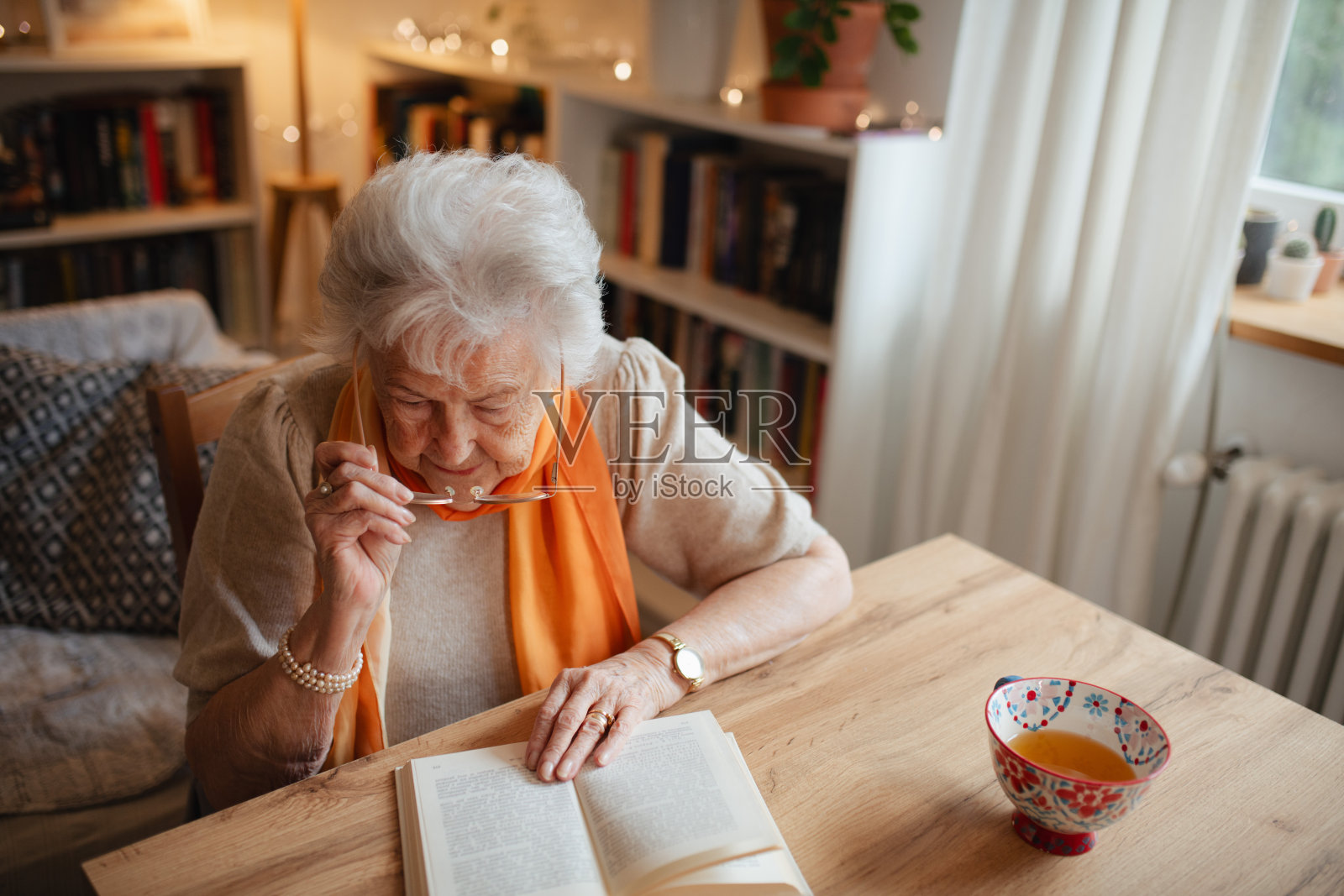可爱的老奶奶坐在厨房桌旁喝茶，正在阅读一本书。照片摄影图片
