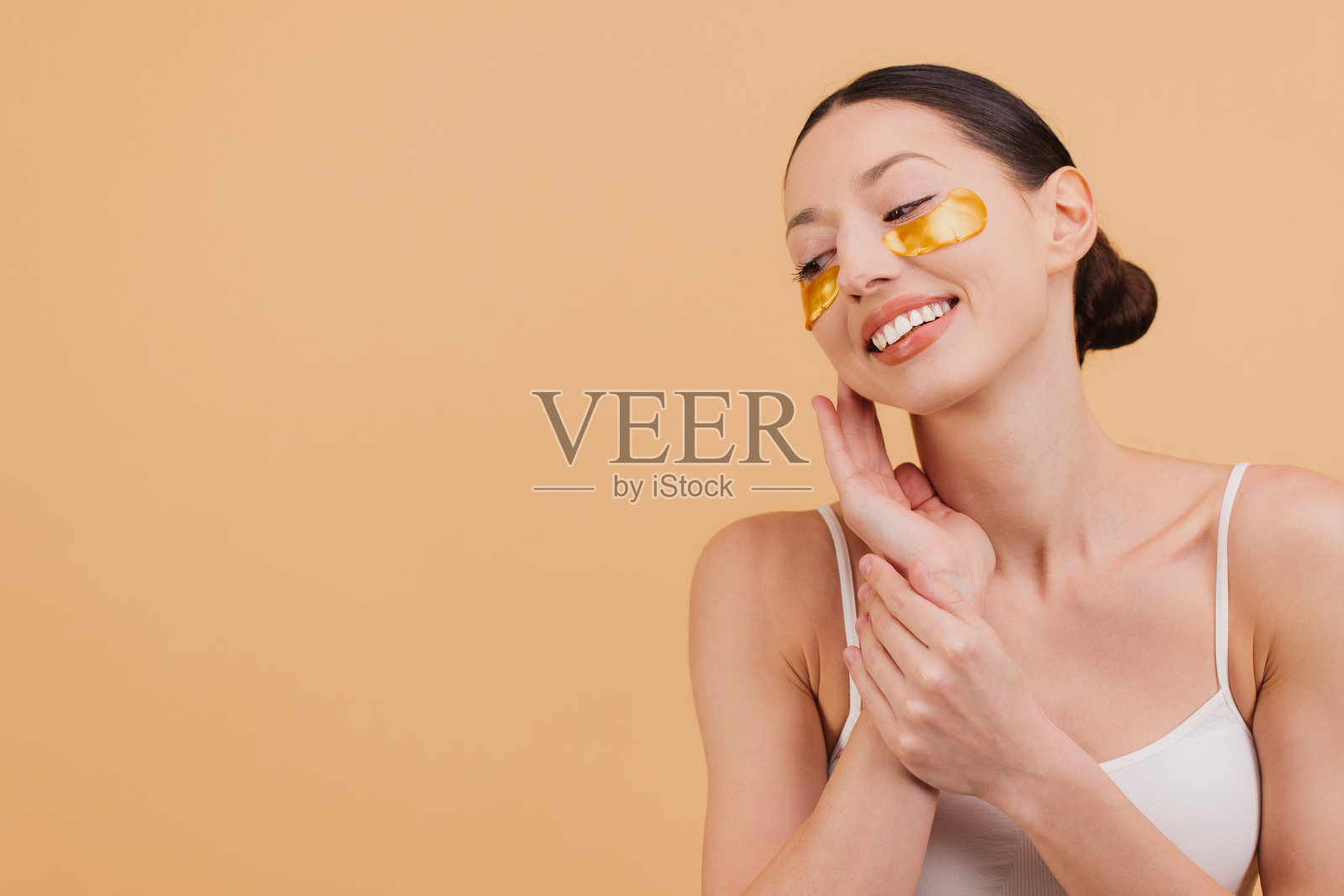 微笑的亚洲女性正在涂抹金色眼贴，轻触她的脸庞。照片摄影图片