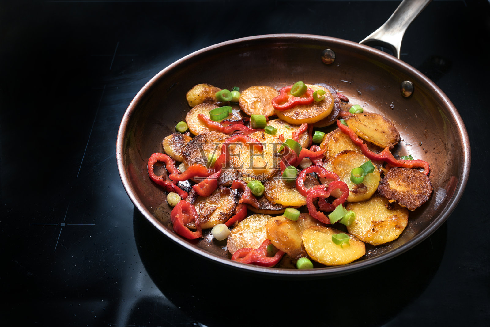 脆炸土豆、红椒和绿色春葱在家里的黑色灶台上的煎锅中，烹饪蔬菜，素食餐，特写焦点照片摄影图片