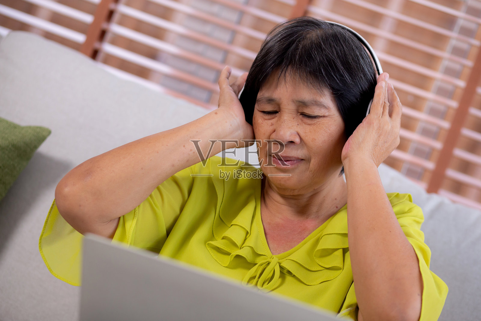 快乐的亚洲老年女性坐在沙发上戴着耳机听音乐，享受家庭客厅的乐趣，幸福的老年女性与娱乐、生活方式和活动。照片摄影图片