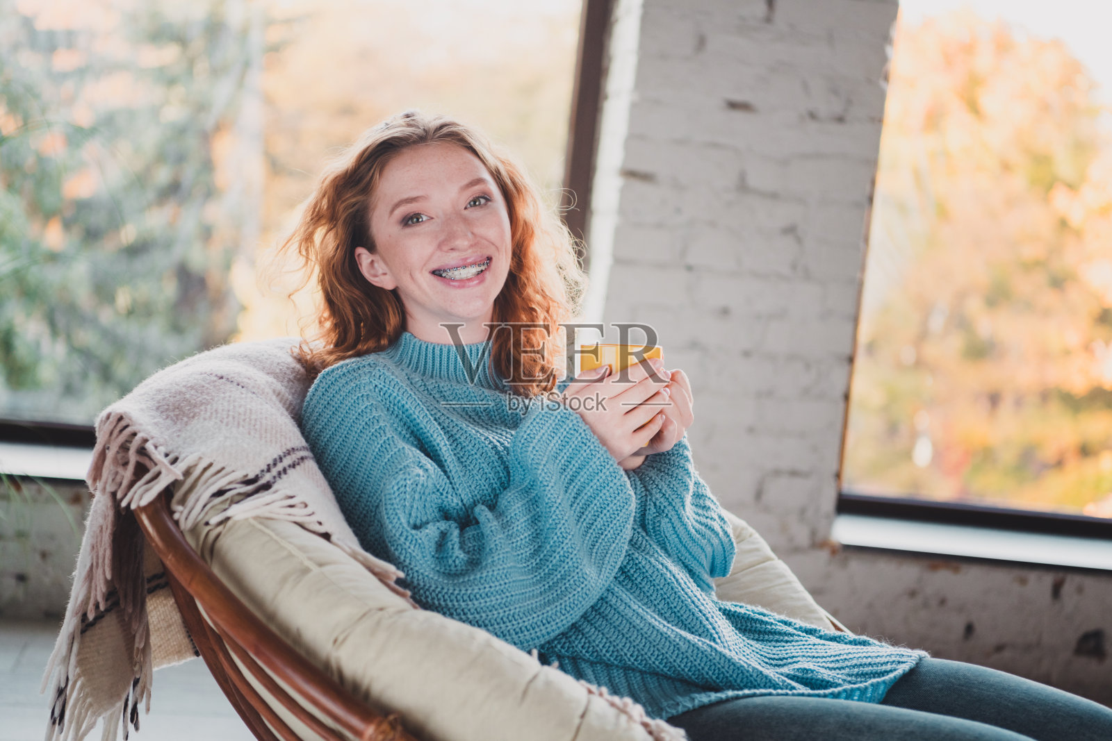 年轻女性在舒适的客厅里，穿着蓝色针织毛衣，手里拿着一个黄色的杯子，沐浴在自然光线中。照片摄影图片