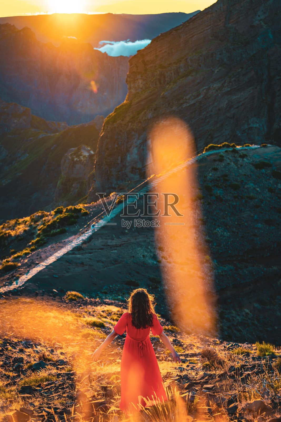 穿着红色连衣裙的美丽女性在阿里埃罗峰享受阳光明媚的傍晚氛围，双臂张开。马德拉岛，葡萄牙，欧洲。照片摄影图片