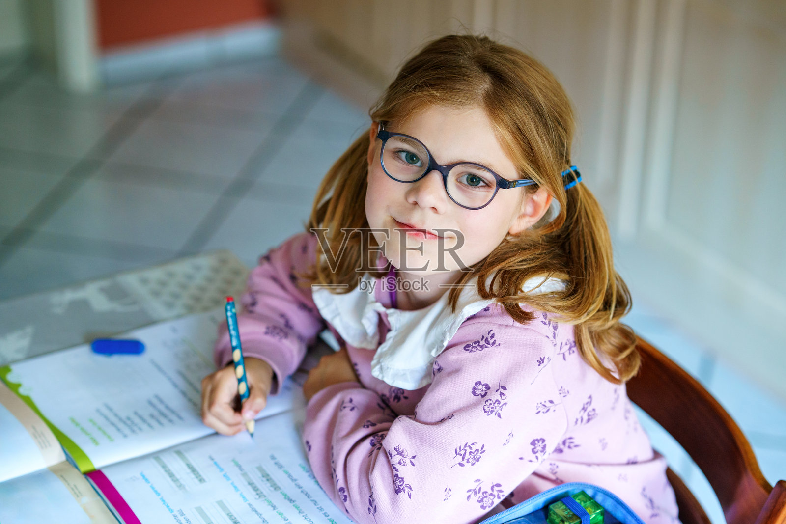 戴眼镜的小女孩穿着粉色毛衣，坐在桌子旁做作业。孩子抬头微笑，手里拿着铅笔，面前放着一本打开的书。室内学习，专注而投入。照片摄影图片
