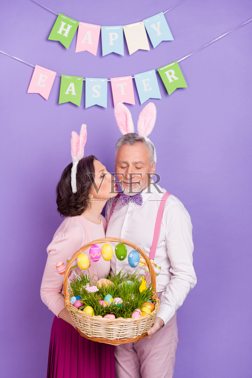 两位年长的和平伴侣在紫色背景下亲吻脸颊，手持装有鸡蛋的食物篮。照片摄影图片