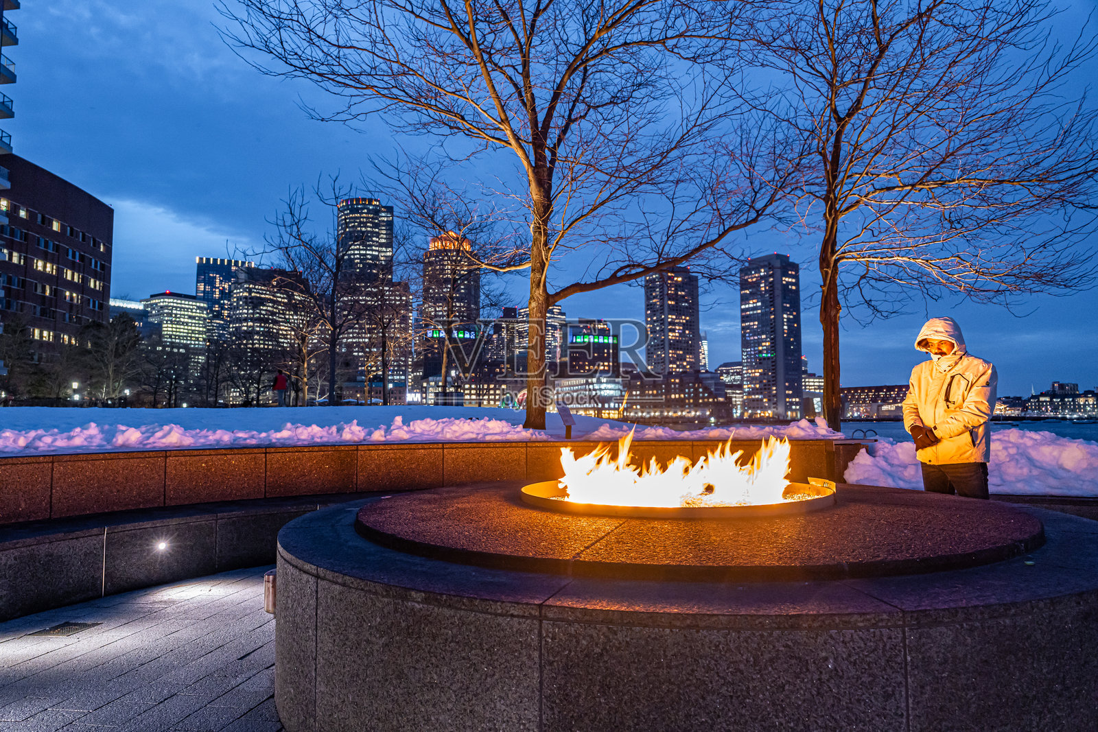 一个男人站在户外火坑旁，火焰熊熊燃烧，背景是波士顿市区的夜空。照片摄影图片