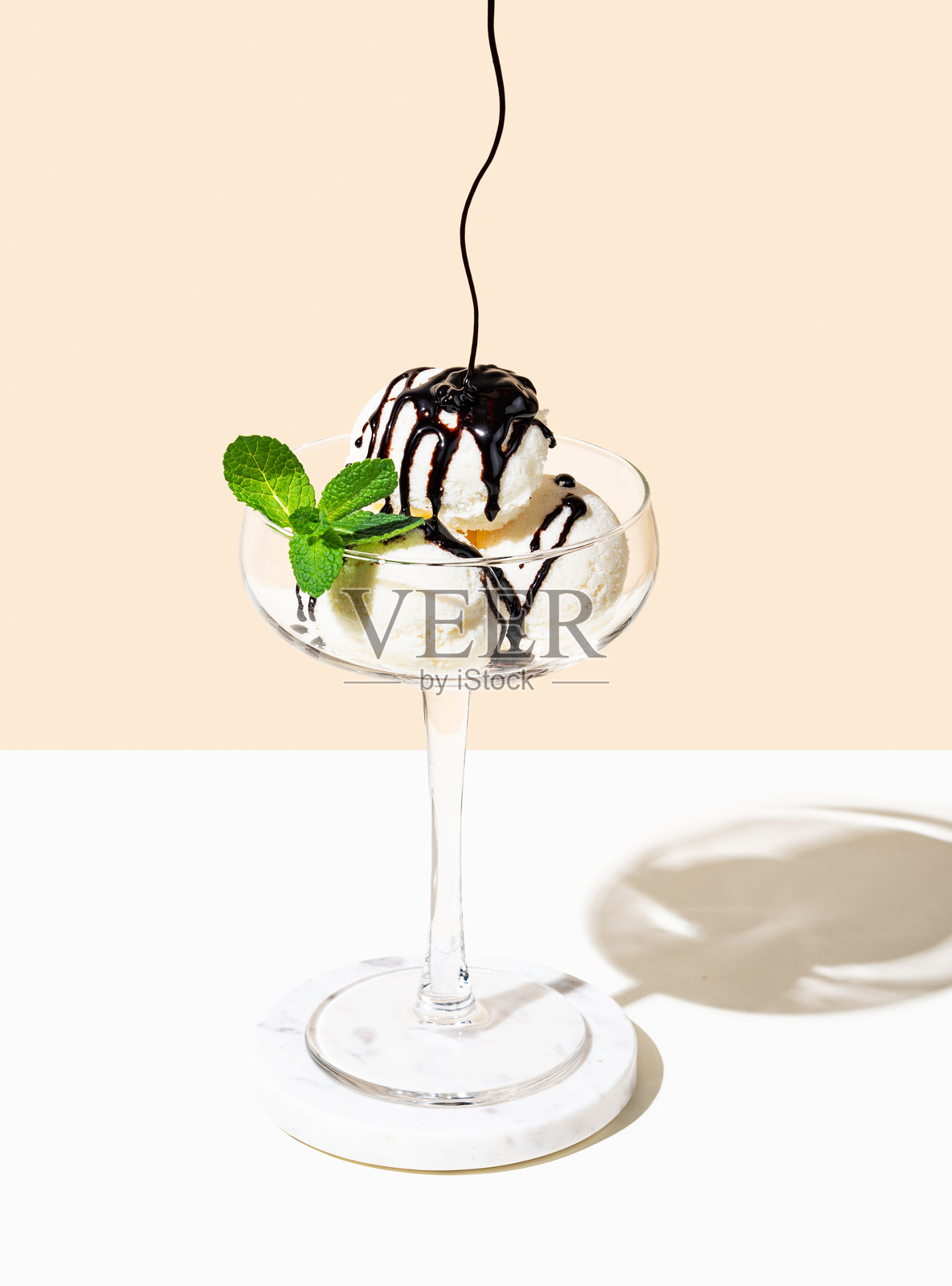 高脚杯中盛满香草冰淇淋，上面淋有巧克力酱，点缀着薄荷叶，背景为浅色。照片摄影图片
