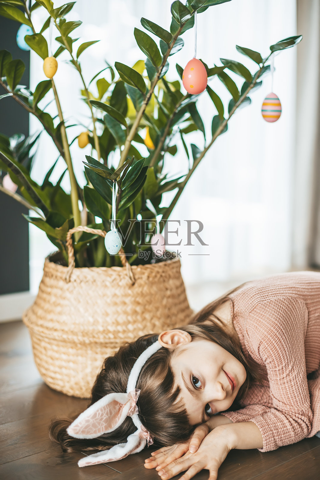 一个穿着粉色裙子、戴着毛茸茸兔耳发箍的微笑女孩，躺在装饰有节日复活节彩蛋的室内植物前。照片摄影图片