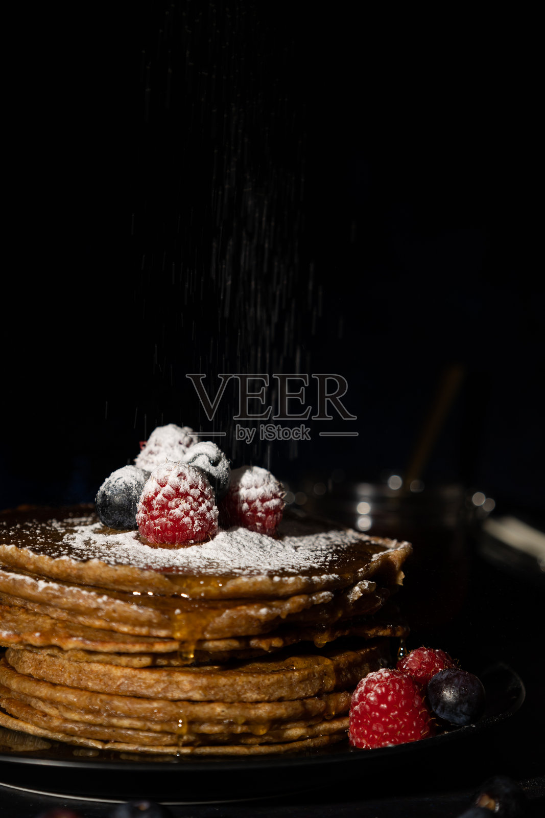 垂直图像展示了叠放的煎饼，上面撒有糖粉，点缀着红色覆盆子和蓝莓，放在黑色盘子上，背景为深色。照片摄影图片