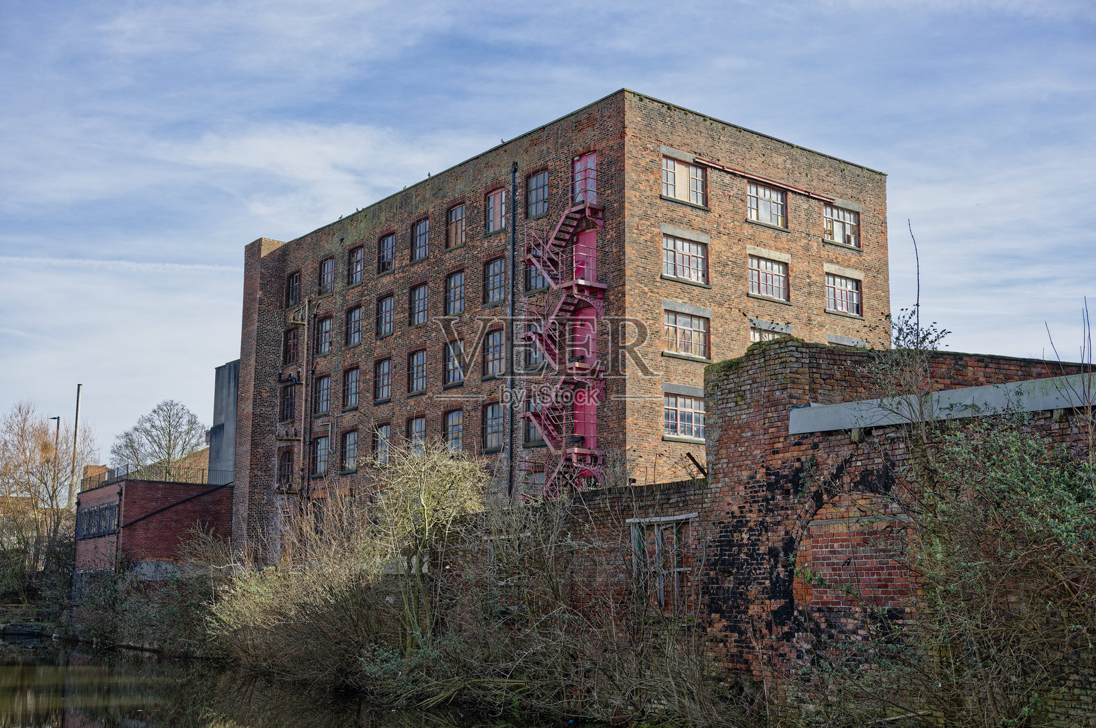 历史悠久的砖厂磨坊建筑，旁边有红色消防梯，靠近水边，天气晴朗。照片摄影图片