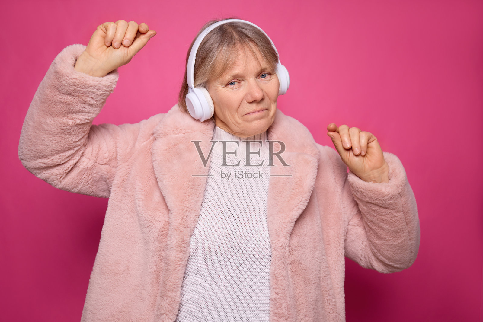 一位快乐的女性穿着舒适的粉色外套和白色耳机，随着音乐跳舞，展现出她无忧无虑的精神，背景是充满活力的粉色。照片摄影图片