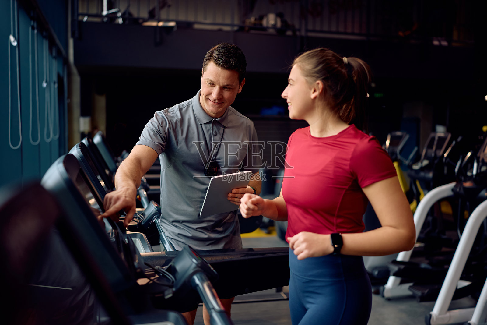 快乐的私人教练在健身房监测运动女性在跑步机上的跑步。照片摄影图片