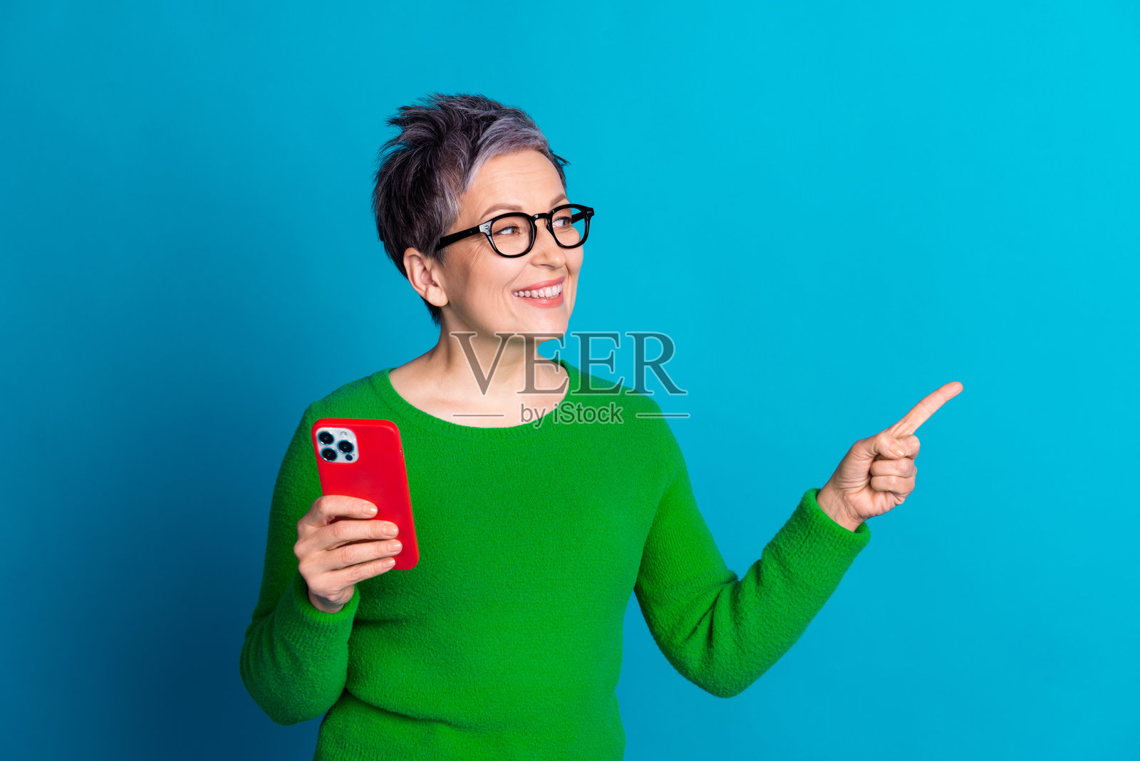 穿着绿色毛衣的快乐老年女性微笑着，手持智能手机并指向鲜艳的蓝色背景。照片摄影图片
