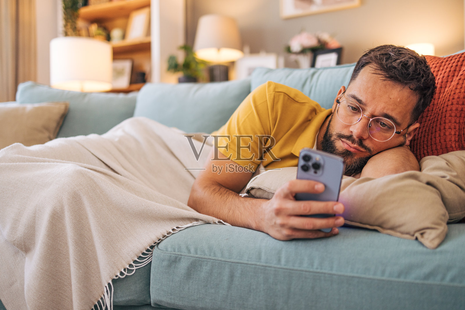 一个中年悲伤的男人躺在客厅的沙发上，正在使用他的智能手机。照片摄影图片