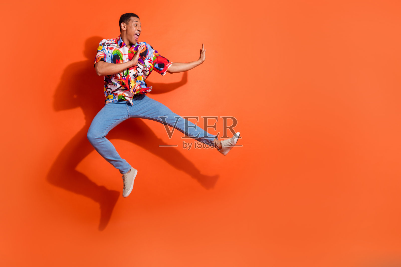 快乐的男人在充满活力的橙色背景下充满活力地跳跃，穿着印花衬衫和休闲牛仔裤，体现时尚潮流照片摄影图片