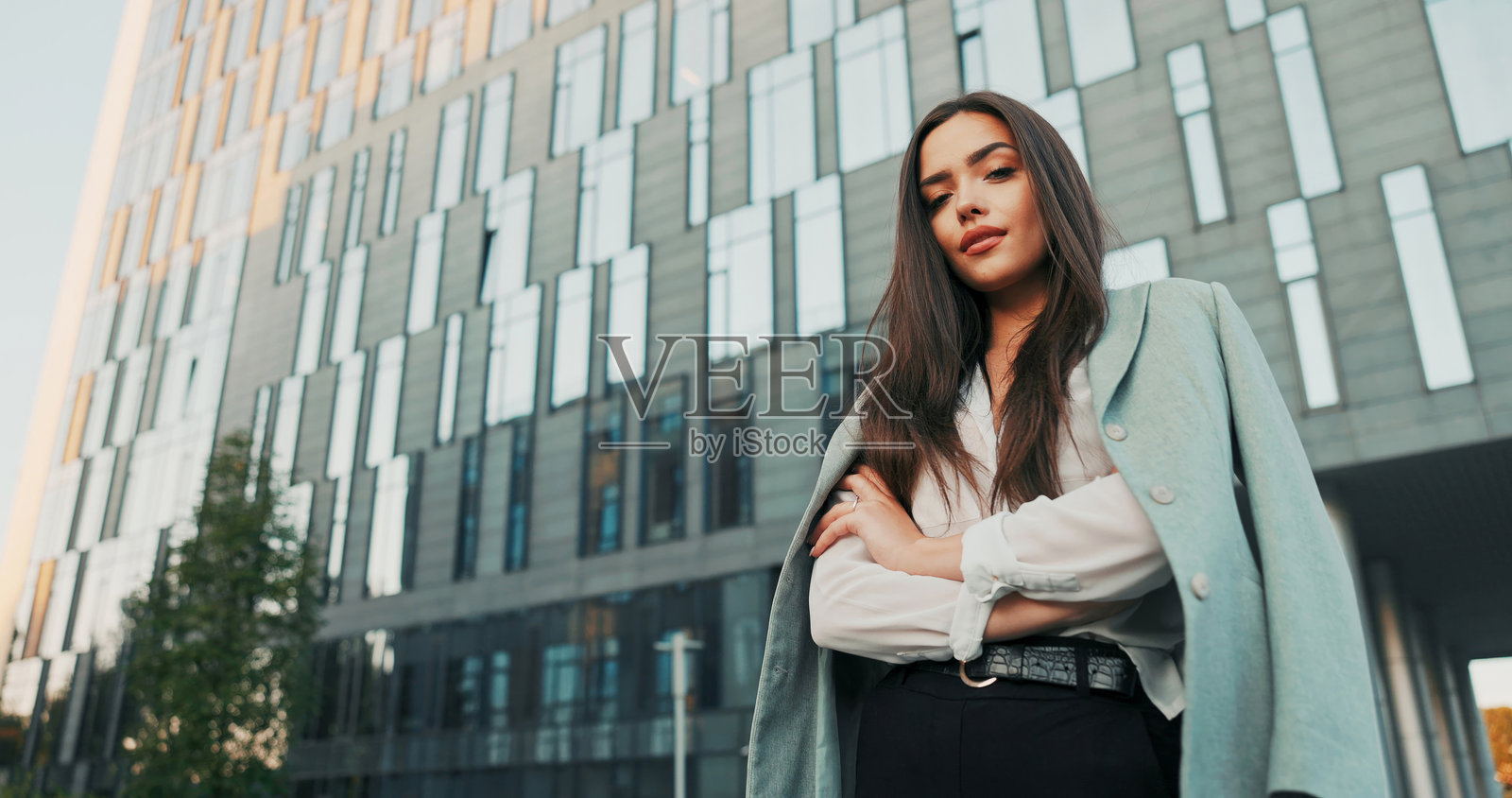 一位身材出众的雄心勃勃的女性自信地站在一家企业办公室外，穿着时尚的商务服装和高跟鞋，双臂交叉，散发出权威感。照片摄影图片