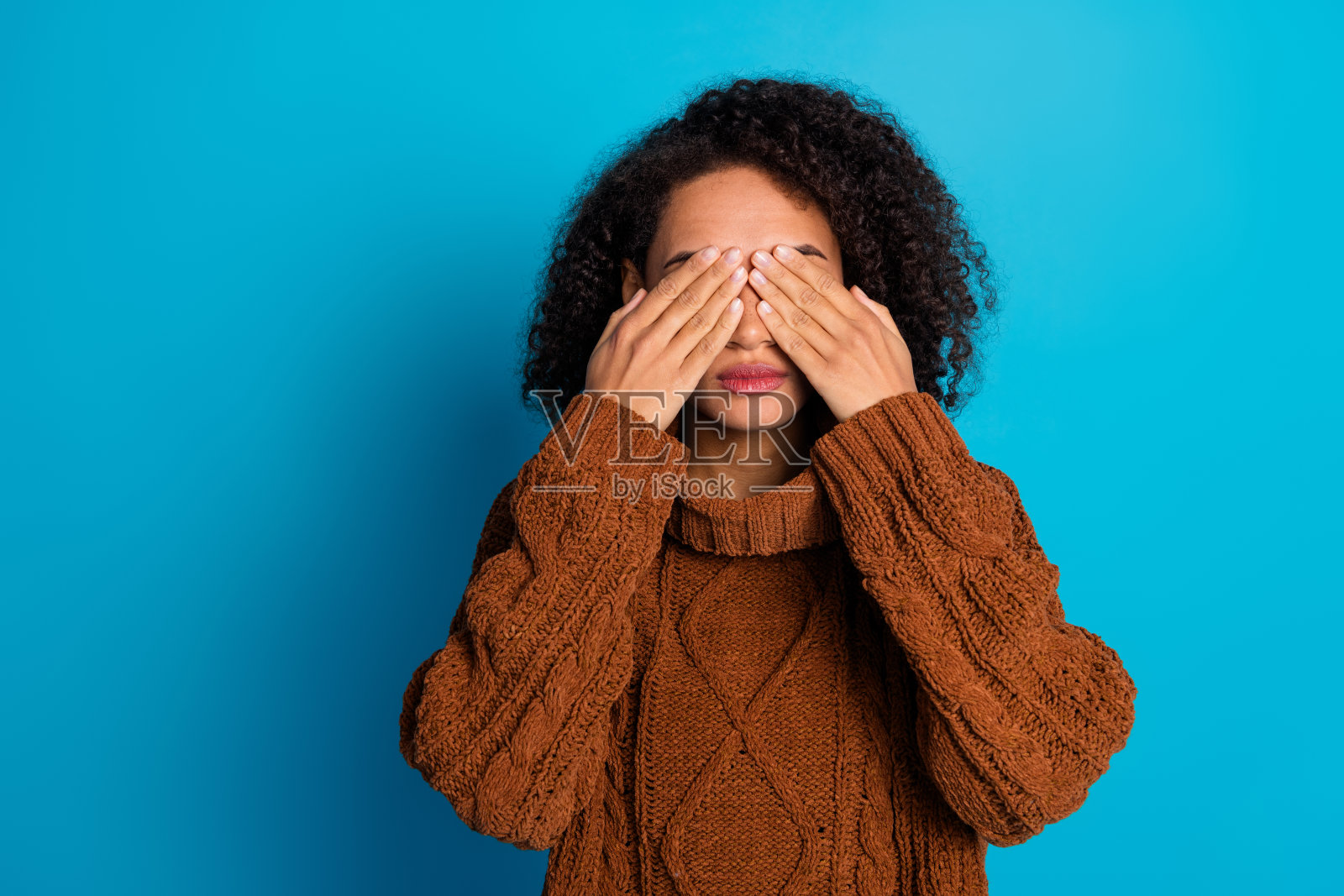 时尚年轻女性用手遮住眼睛，穿着舒适的棕色针织毛衣，背景是鲜艳的蓝色。照片摄影图片
