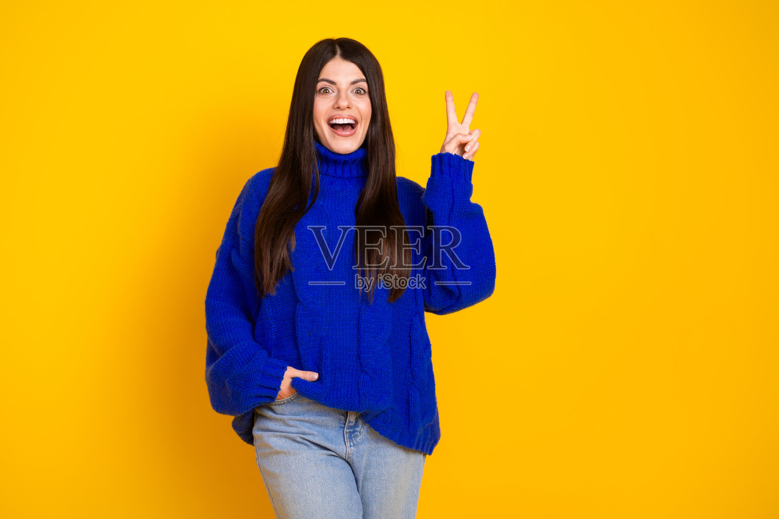 微笑的女性在明亮的黄色背景下，穿着现代蓝色毛衣，做出和平手势。照片摄影图片