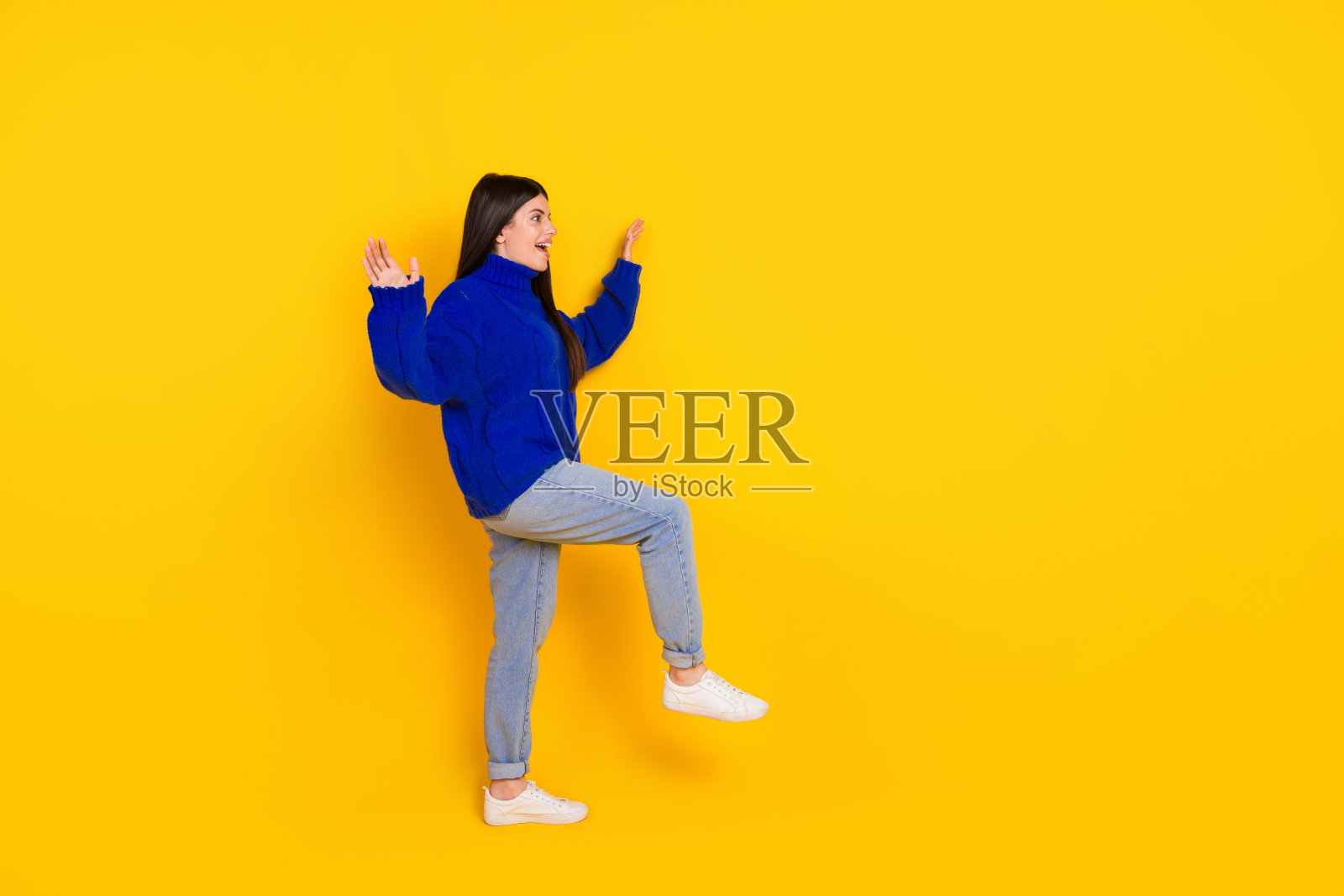 穿着蓝色毛衣的快乐年轻女性在鲜艳的黄色背景前俏皮地做手势照片摄影图片