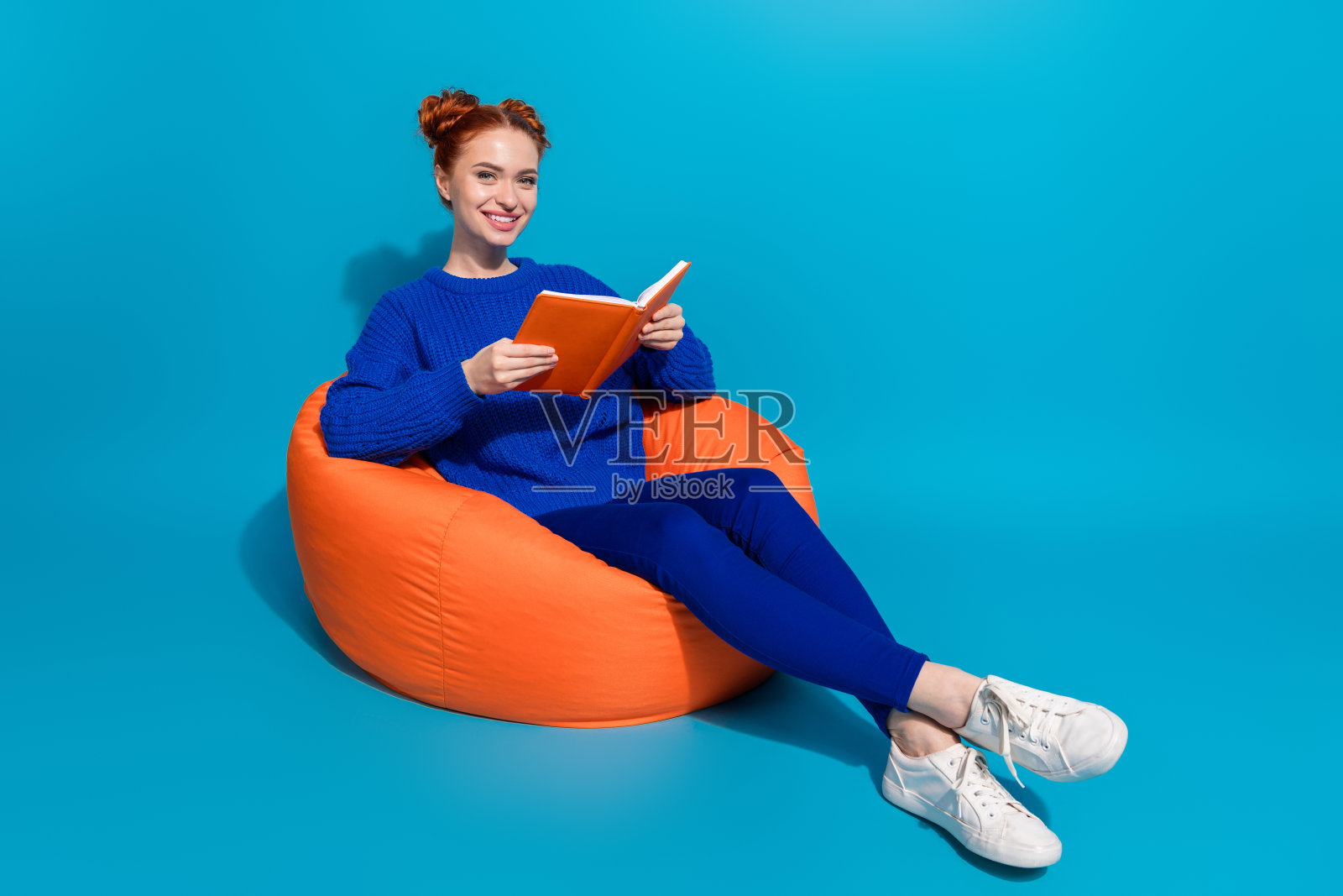 年轻女性在明亮的蓝色背景下，坐在豆袋椅上放松地阅读。照片摄影图片