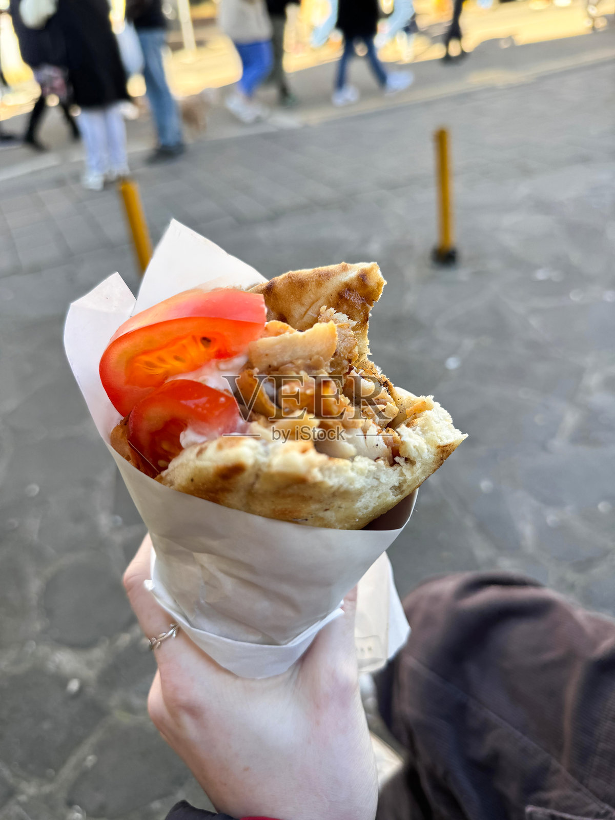 特写镜头中，一位无法辨认的女性手持用防油纸包裹的鸡肉和番茄串，街头美食，户外市场背景，焦点在前景。照片摄影图片