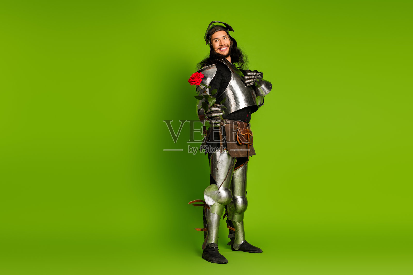 年轻的中世纪骑士手持玫瑰，身穿全钢盔甲，站在生机勃勃的绿色背景中，象征着骑士精神和浪漫。照片摄影图片