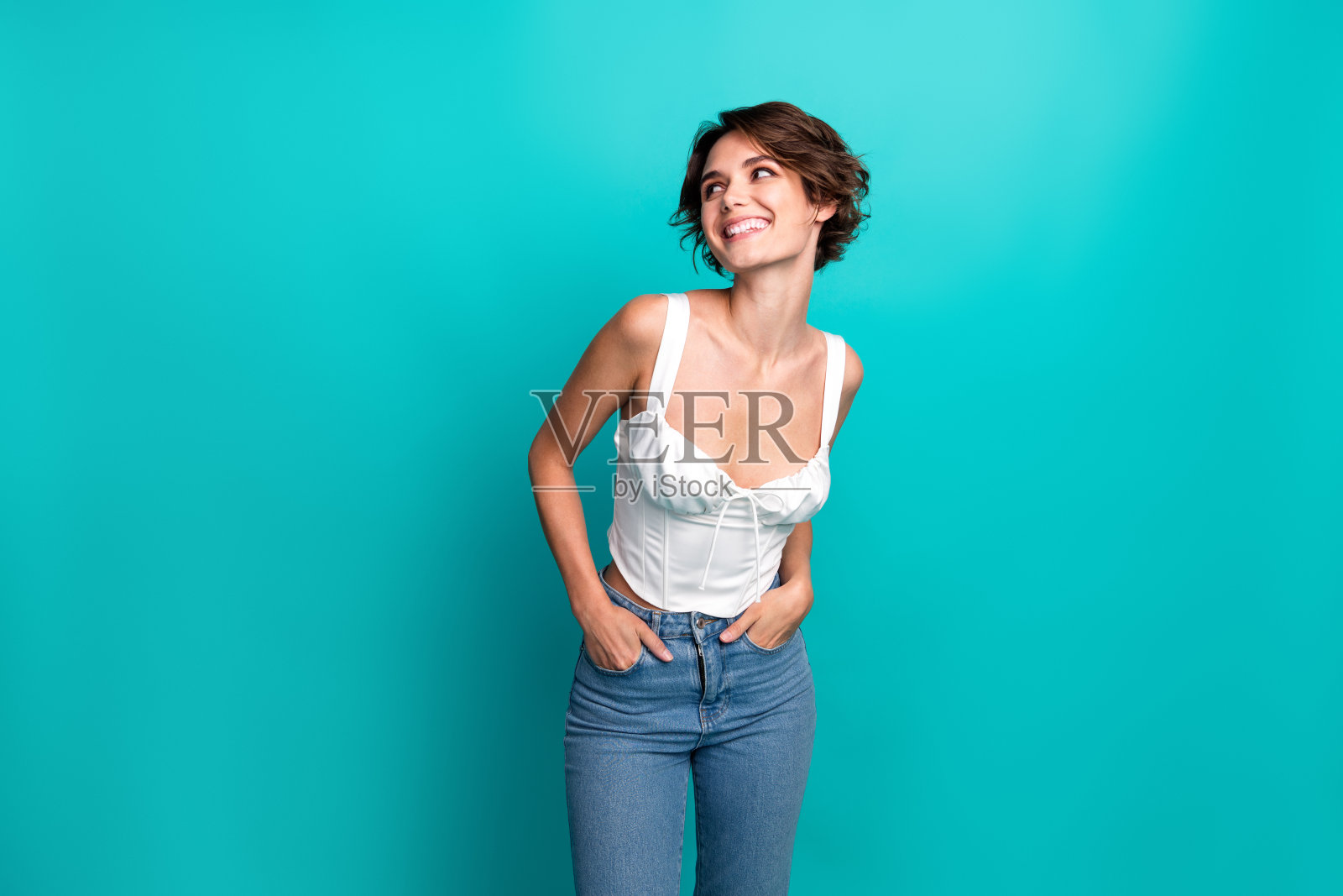 穿着休闲白色上衣和牛仔裤的时尚女性在青绿色背景前微笑，展现出快乐和自信。照片摄影图片
