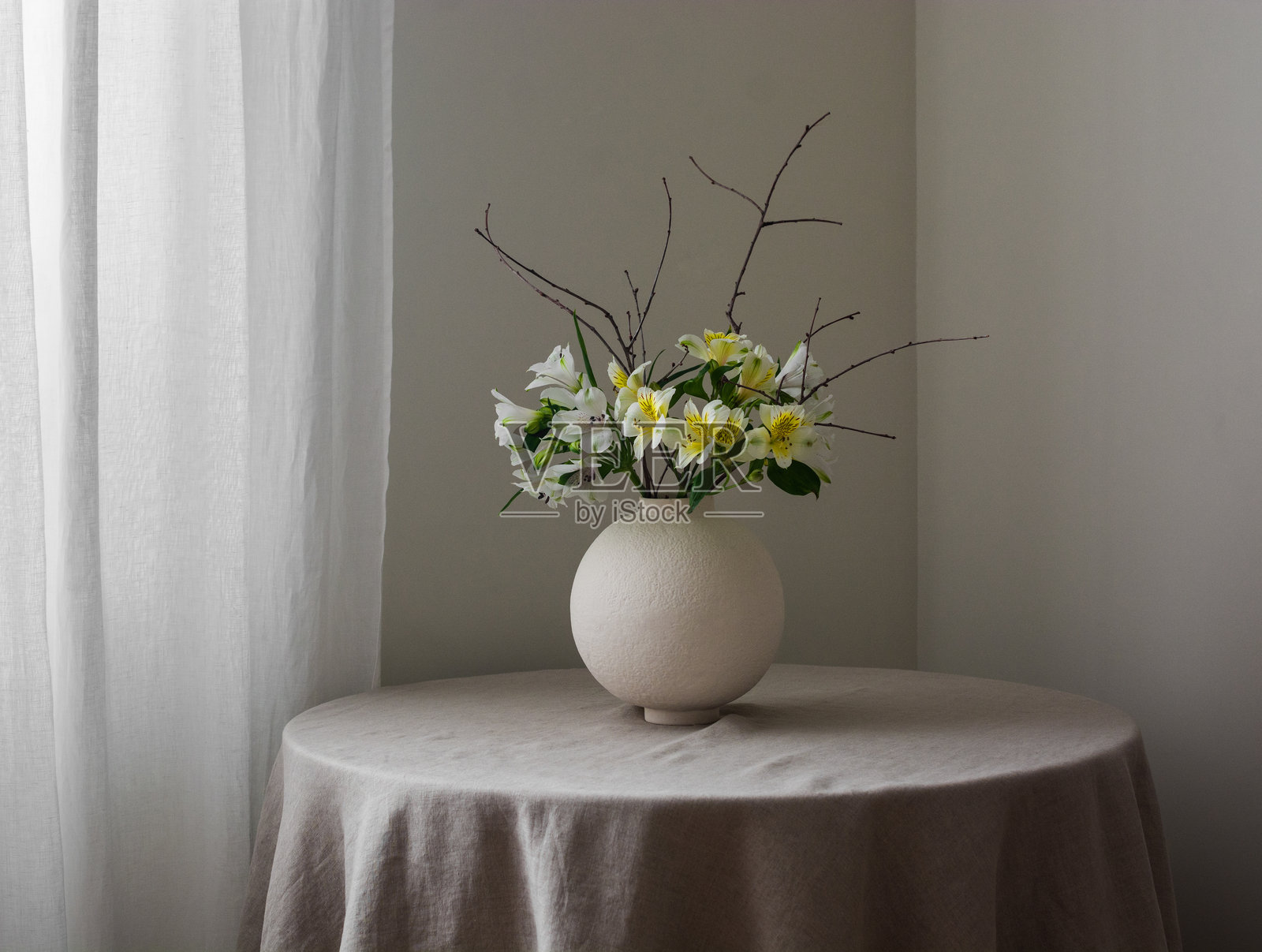 室内装饰的春季花卉布置 - 新鲜的百合花和干枝在圆形陶瓷花瓶中，放置在灰色亚麻桌布上的桌子上照片摄影图片