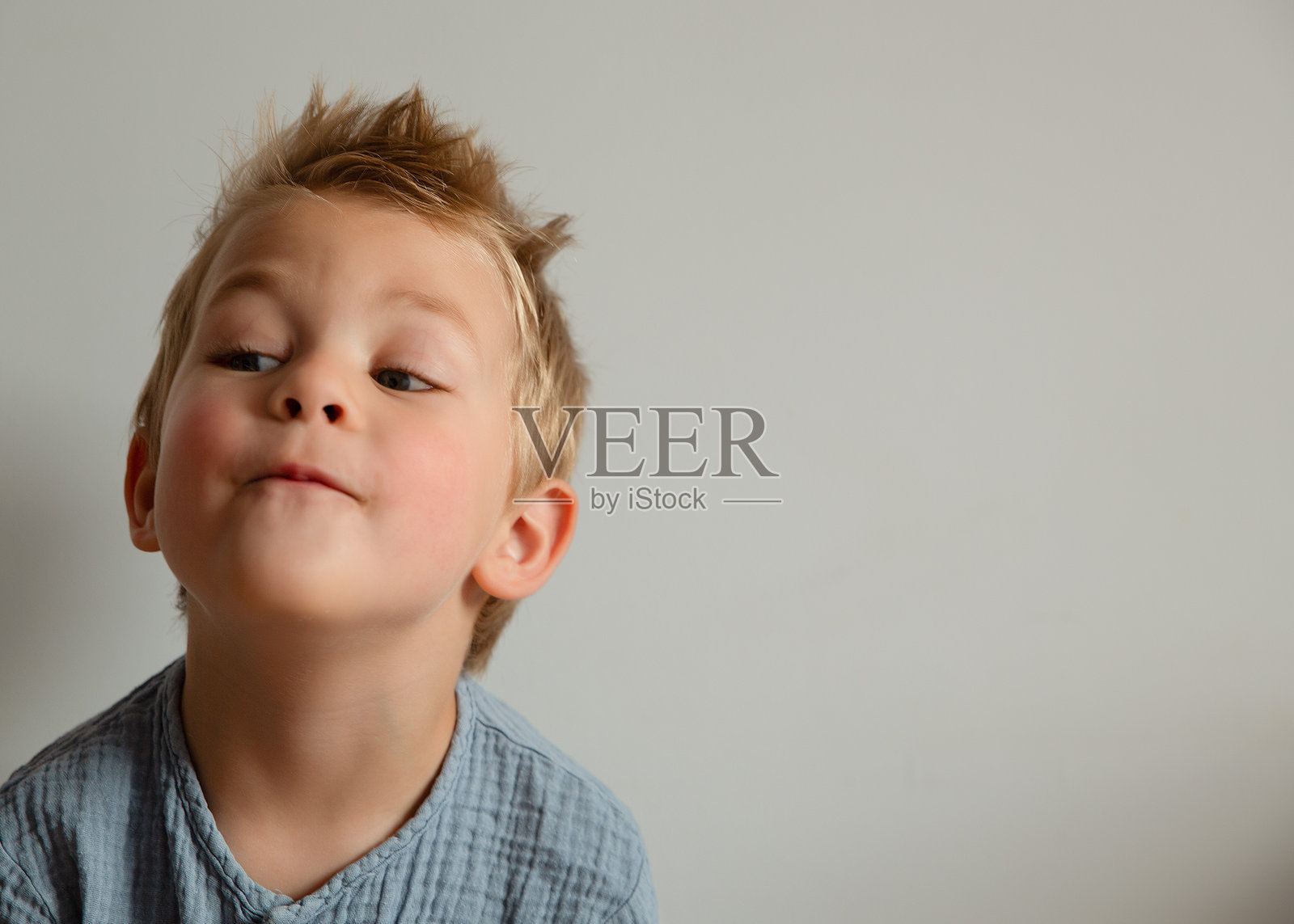 一个面带顽皮微笑、金色乱发、表情调皮的小男孩。这个孩子抬头做着搞笑的表情。背景明亮，摄影棚肖像。照片摄影图片